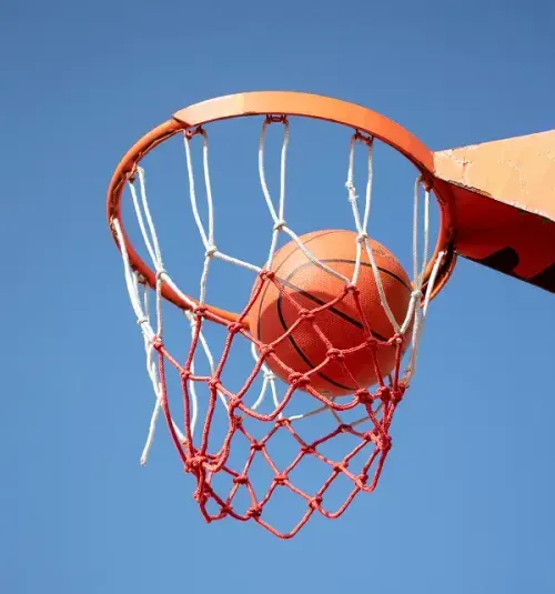 Basketball going through an orange basketball hoop with a red and white net against a clear blue sky.
