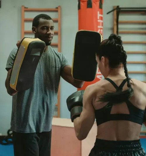 Man holding boxing pads while a woman practices punching in a gym.