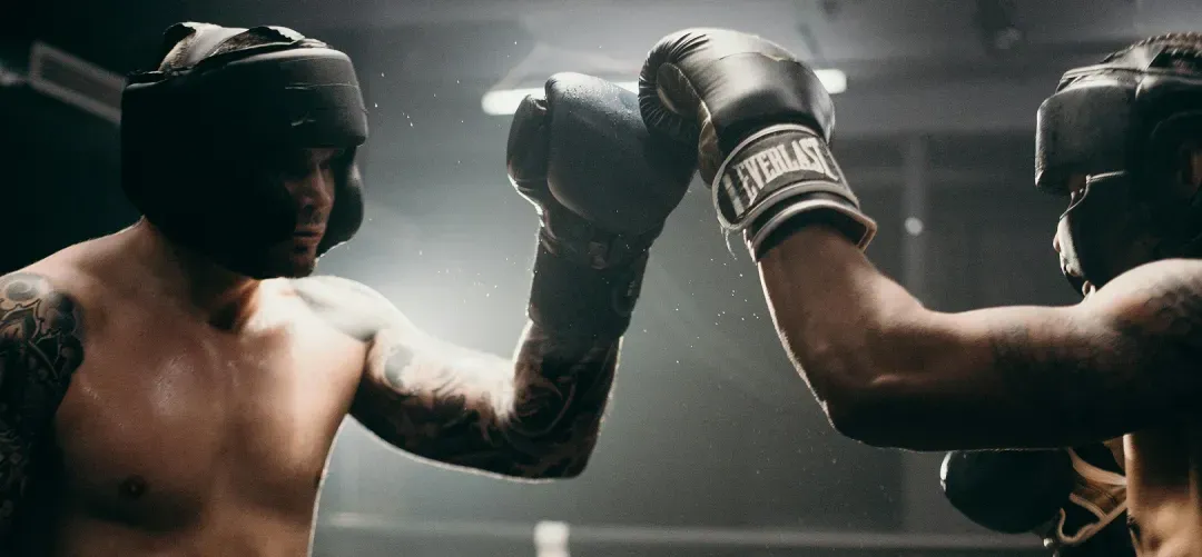Two boxers wearing headgear and gloves sparring in a dimly lit boxing ring.