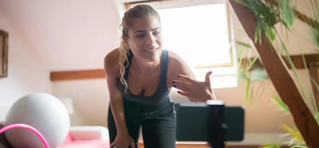 Woman in workout clothes interacting with a smartphone on a tripod in a bright room with exercise equipment and plants.