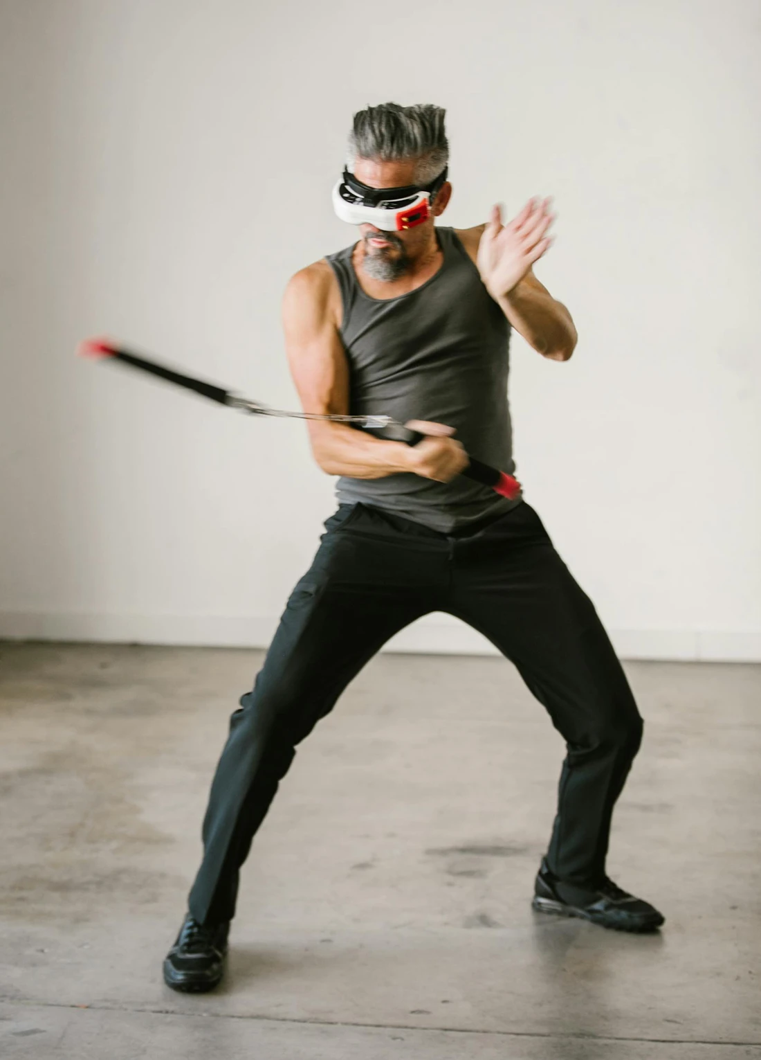 Man in a sleeveless shirt and black pants wearing virtual reality goggles, swinging a motion controller with a dynamic stance.