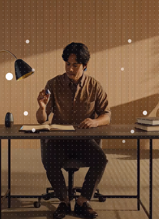 Man in brown shirt sitting at a desk reading a book with a modern black desk lamp and stack of books.