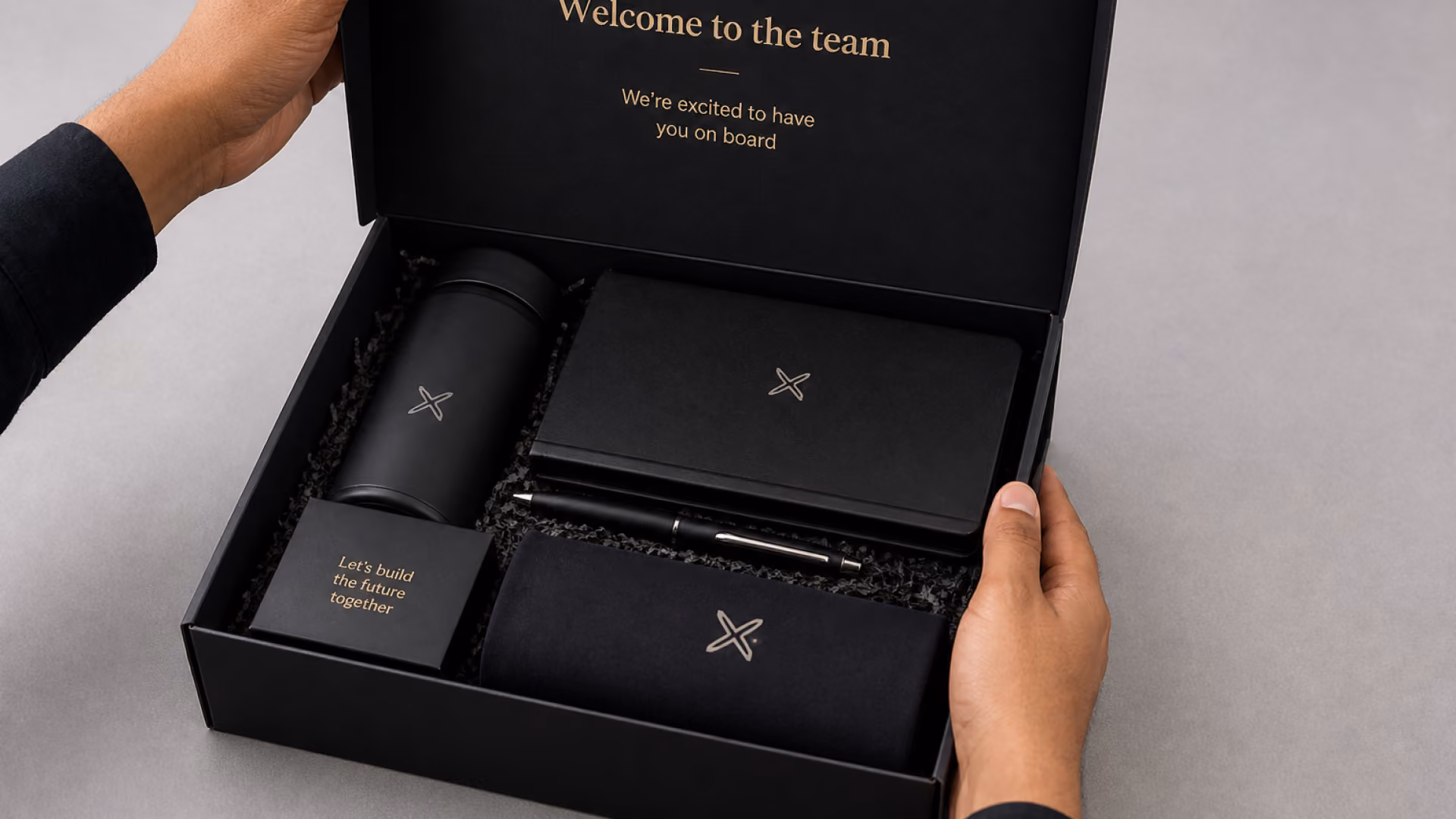 Person opening a luxury corporate onboarding gift box containing branded office essentials, including a notebook, tumbler, pouch, and pen.