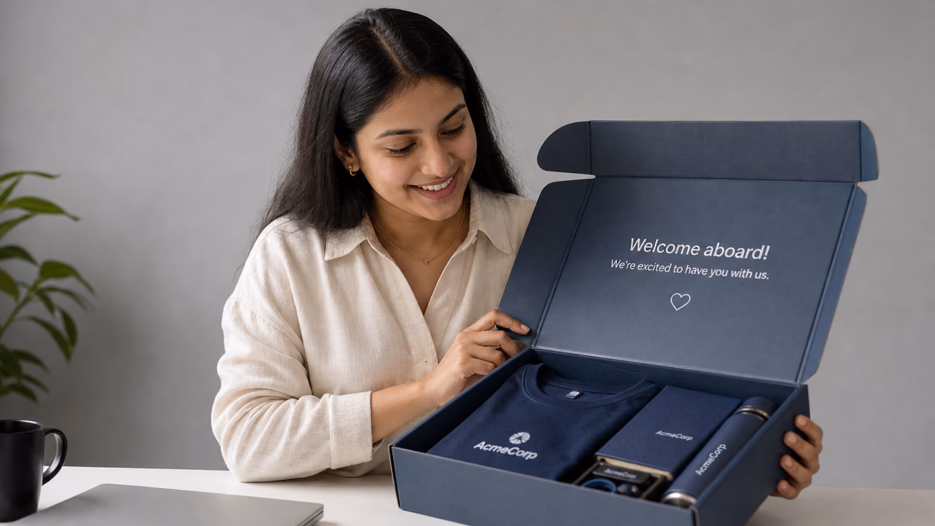 Woman opening a premium custom onboarding gift box containing branded corporate merchandise, including apparel, notebook, and tumbler.