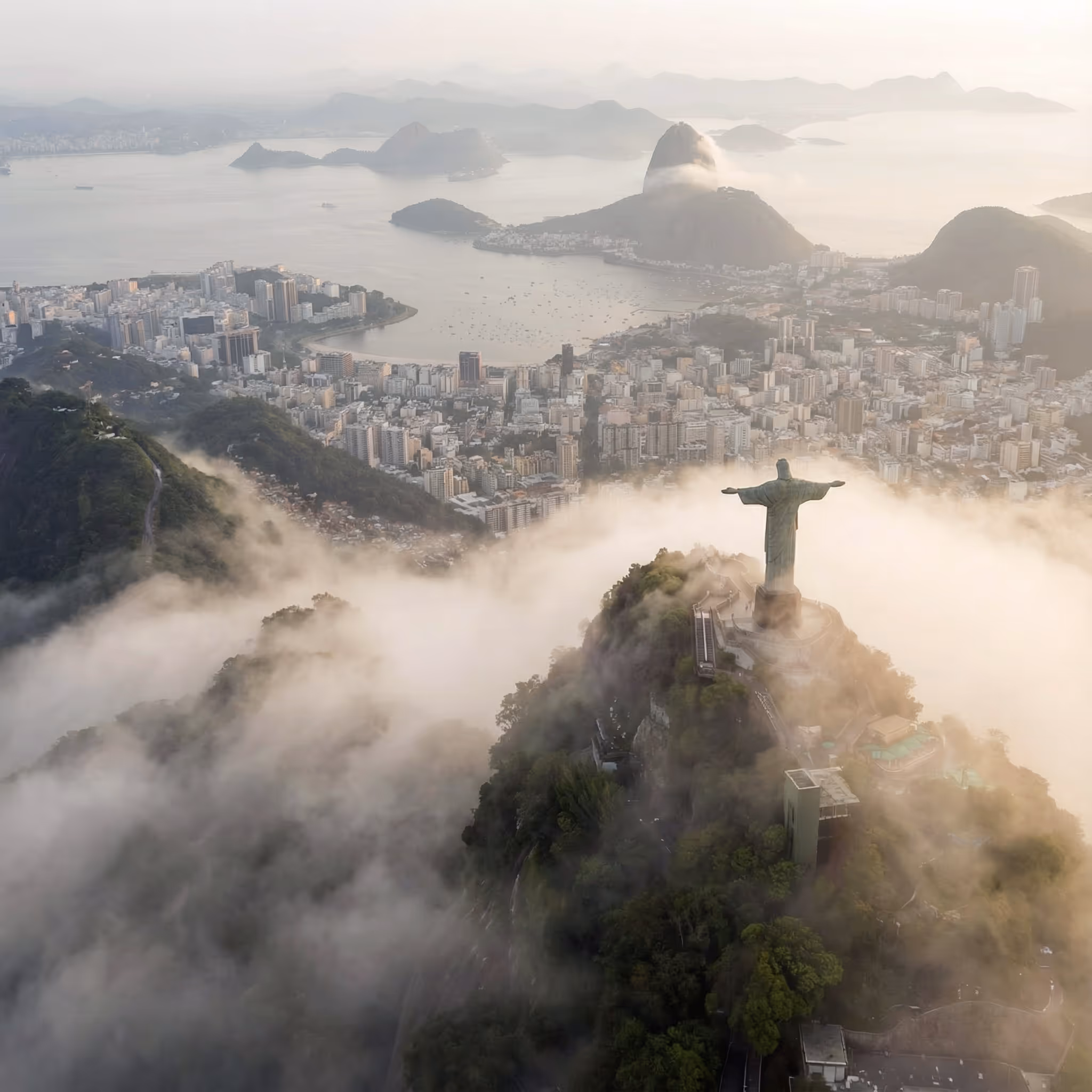 Aerial view of Christ the Redeemer statue surrounded by clouds overlooking Rio de Janeiro with Sugarloaf Mountain in the background.