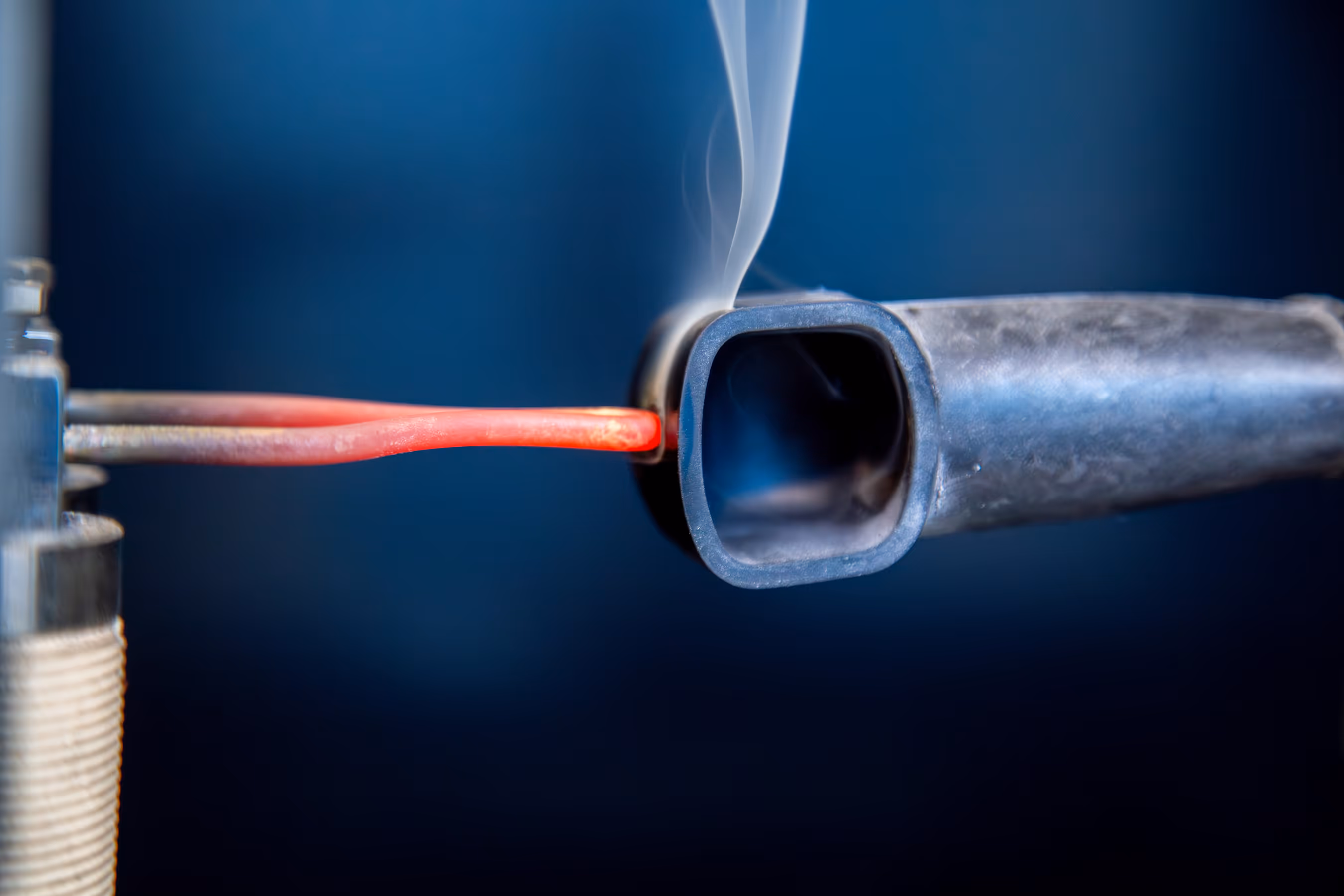 Close-up of a glowing red-hot metal rod inserted into a steel rectangular tube with smoke rising.