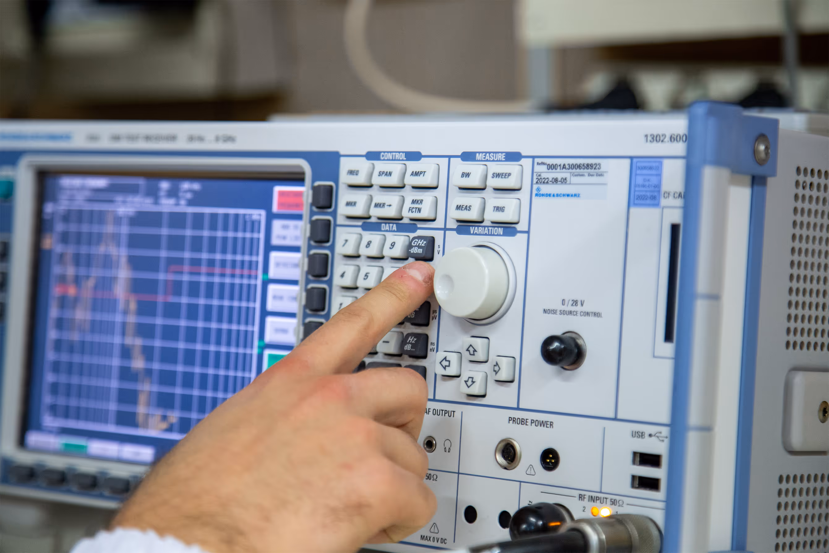 Close-up of a person's hand pressing a button on a signal analyzer showing a frequency spectrum on its screen.