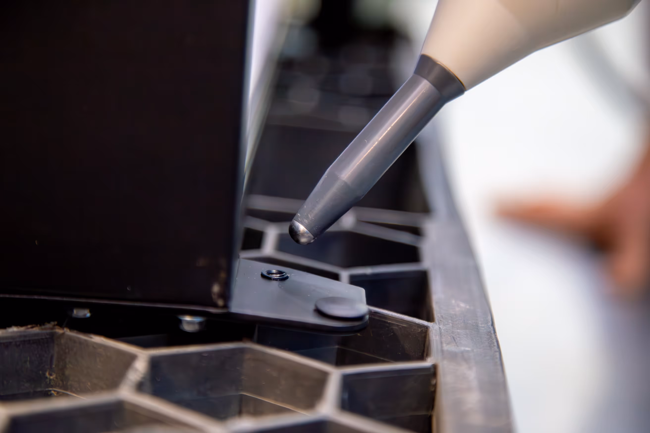 Close-up of a precision measuring probe near a black metal and plastic component with hexagonal pattern.