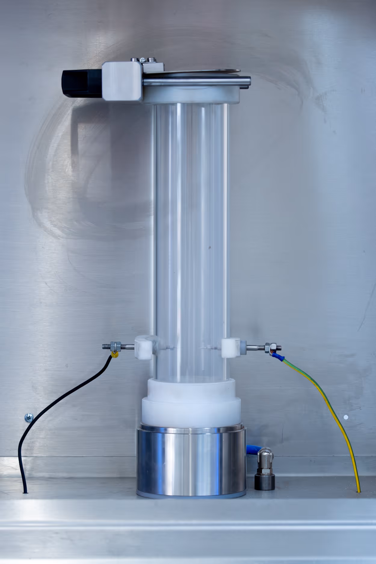 Laboratory apparatus featuring a transparent vertical cylindrical tube with metal and plastic fittings and connected wires inside a stainless steel enclosure.