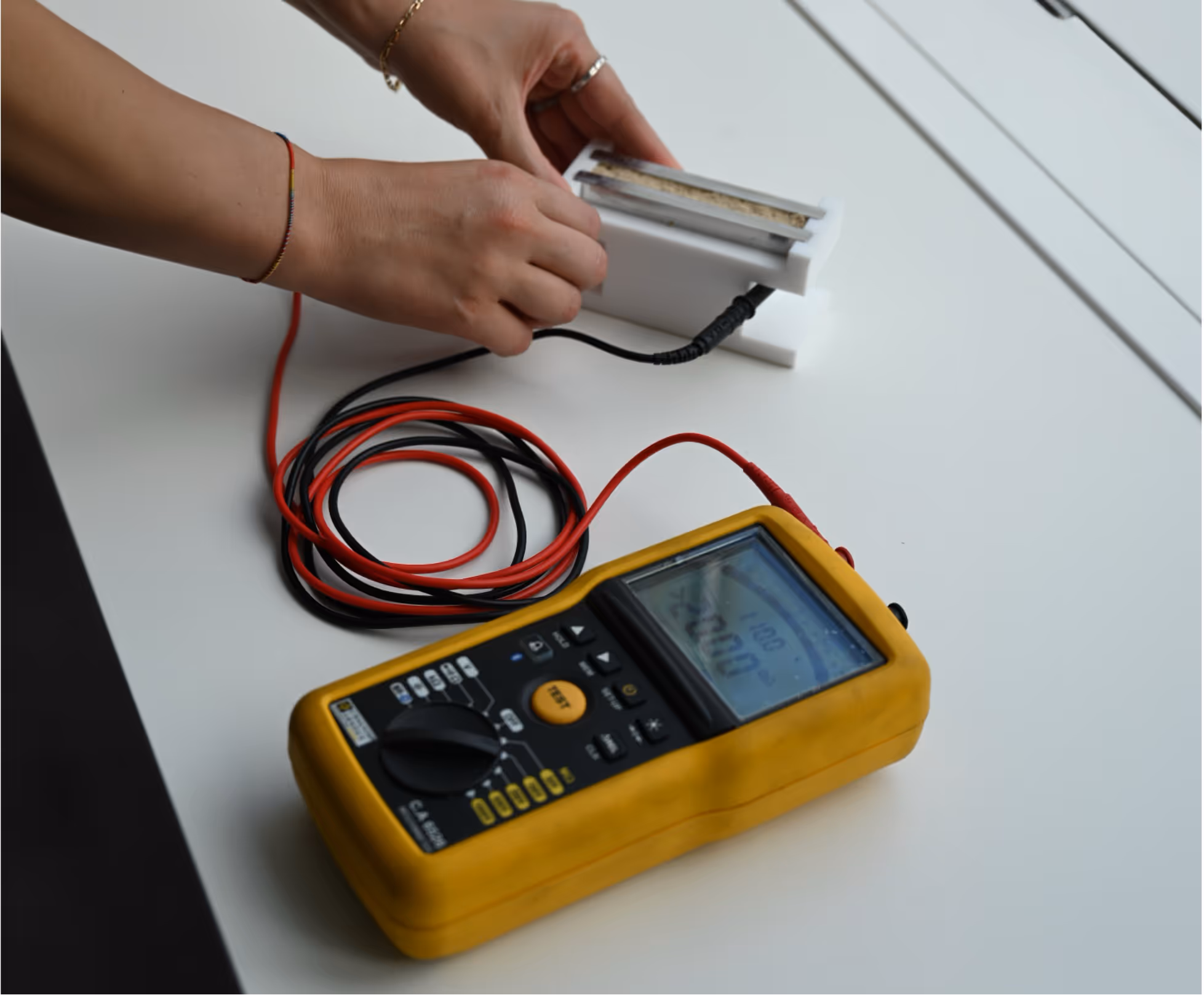 Person holding a white electronic component connected to a yellow digital multimeter with red and black test leads on a white surface.