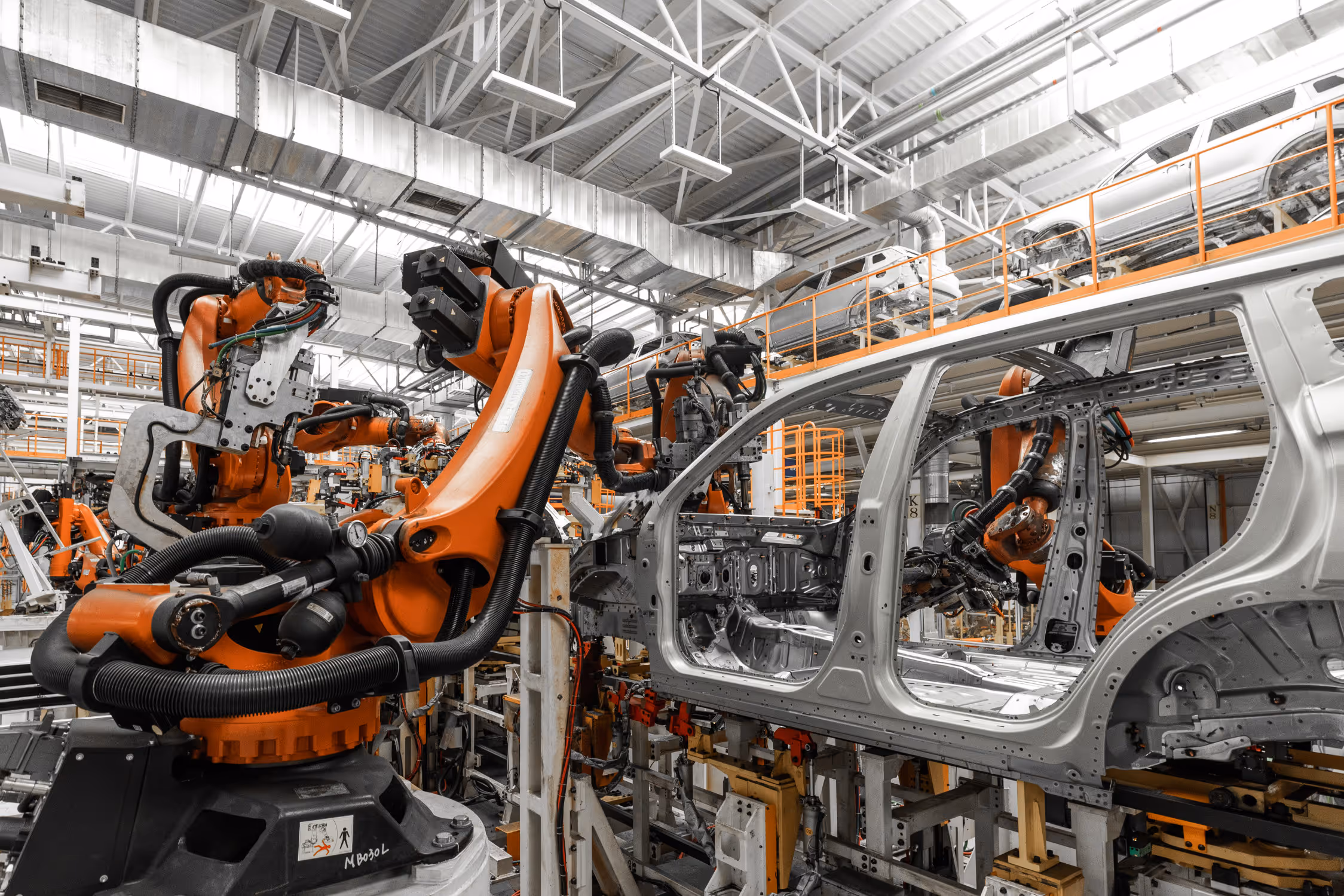 Automated robotic arms assembling a silver car frame inside a large manufacturing factory.