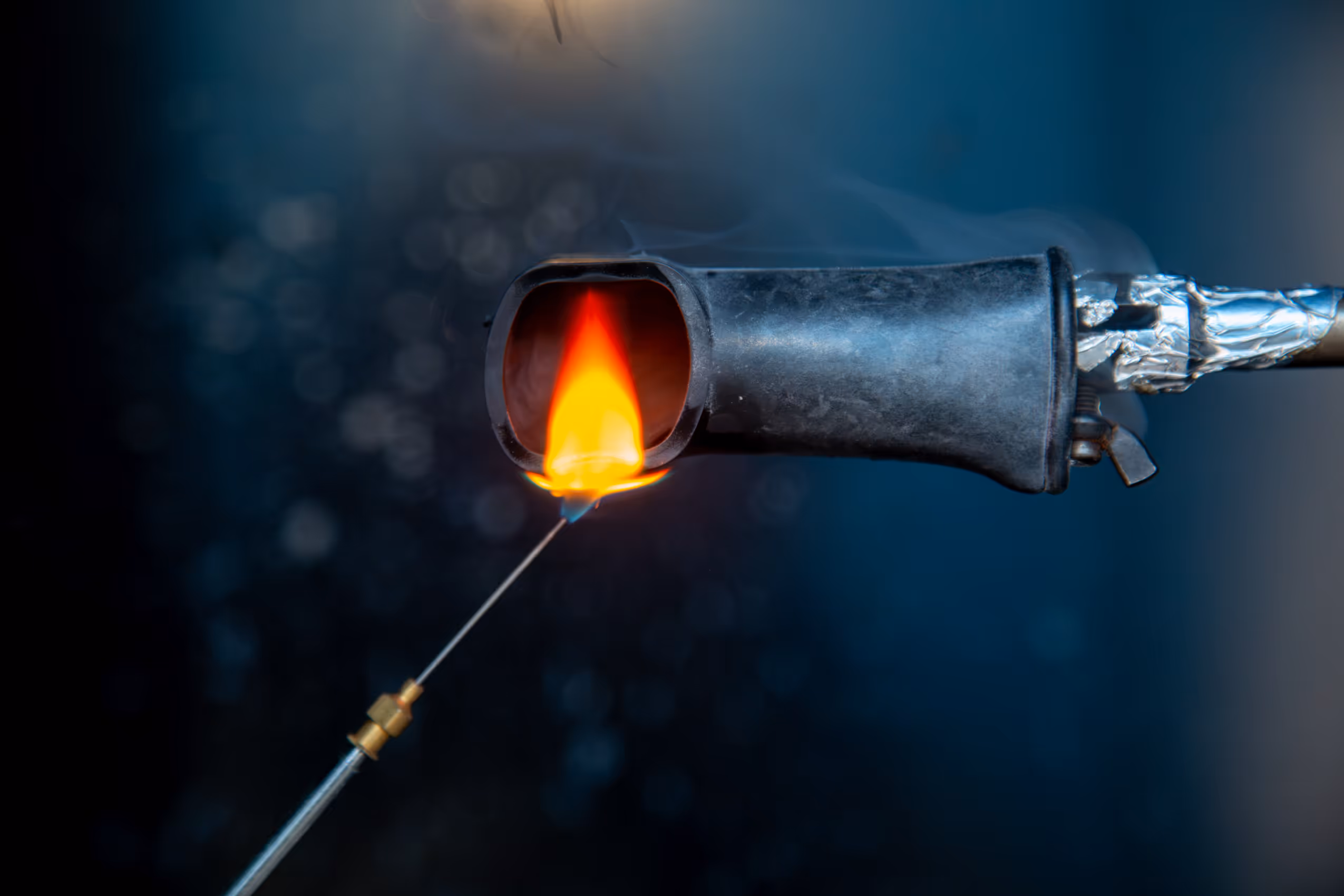 Igniting a flame inside the nozzle of a metal torch with an orange and yellow flame at the tip of a thin rod.