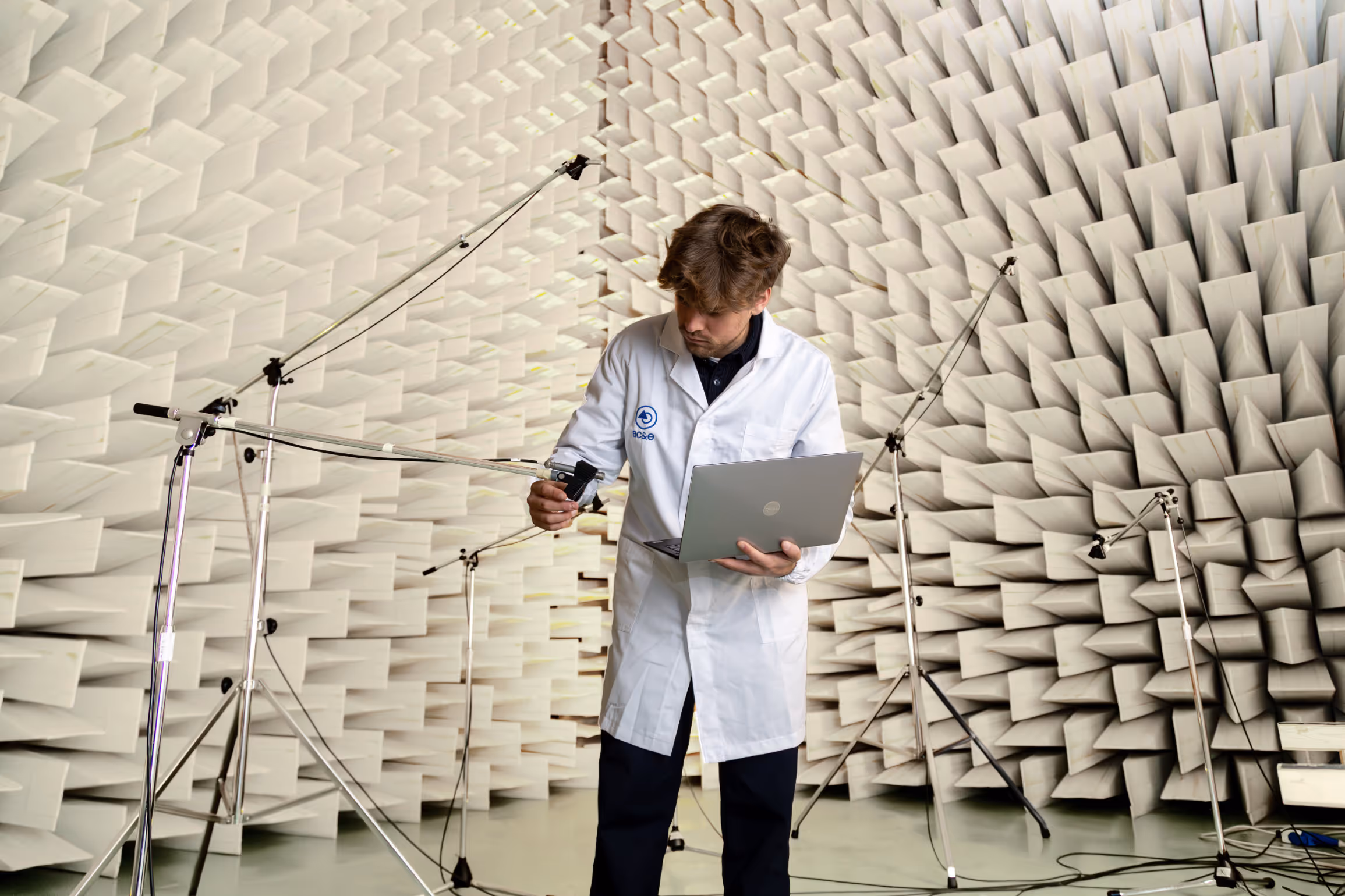 Technician in a white lab coat holding a laptop and adjusting equipment in an anechoic chamber with sound-absorbing panels.