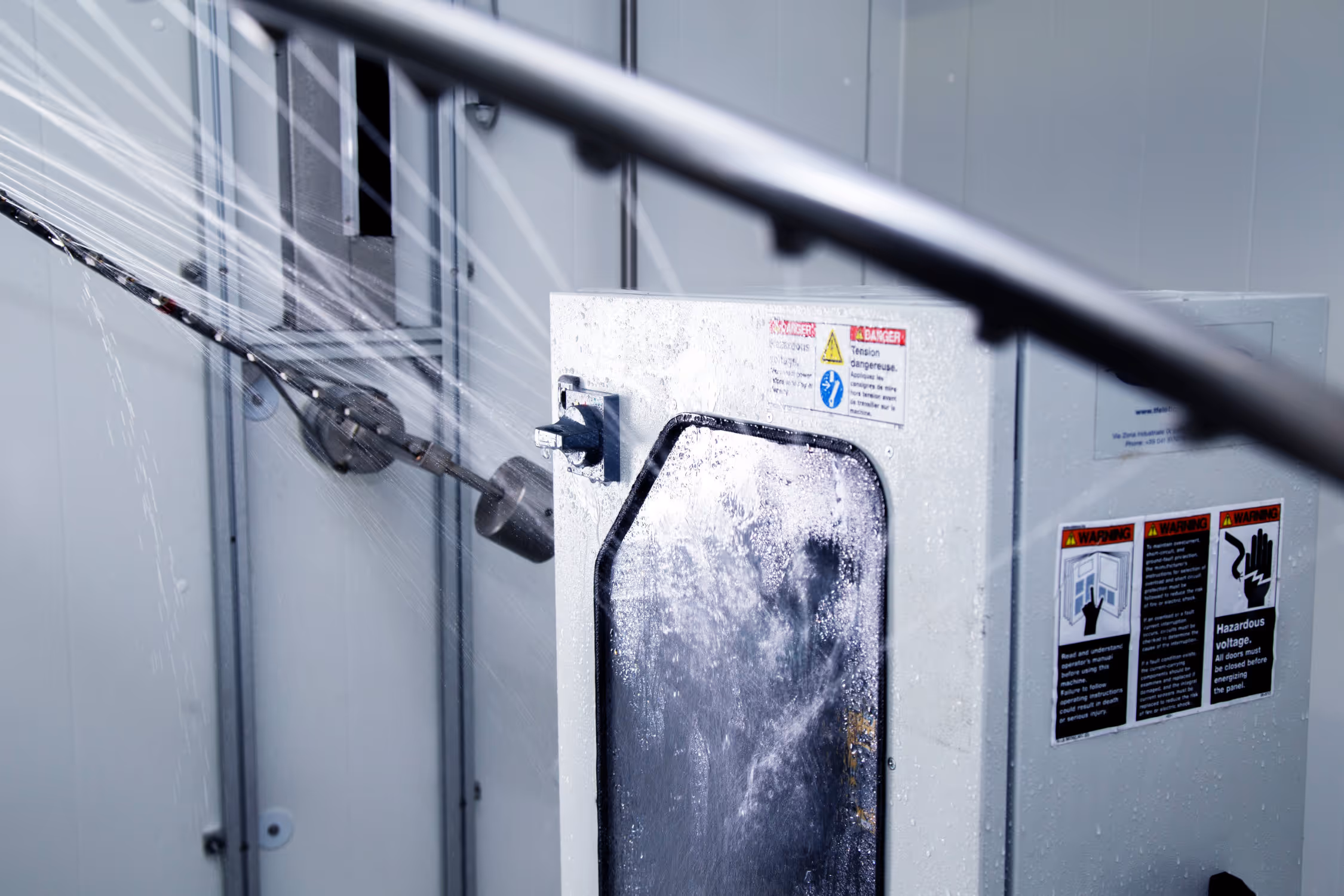 Industrial machine spraying water inside a transparent enclosed compartment with safety warning labels.