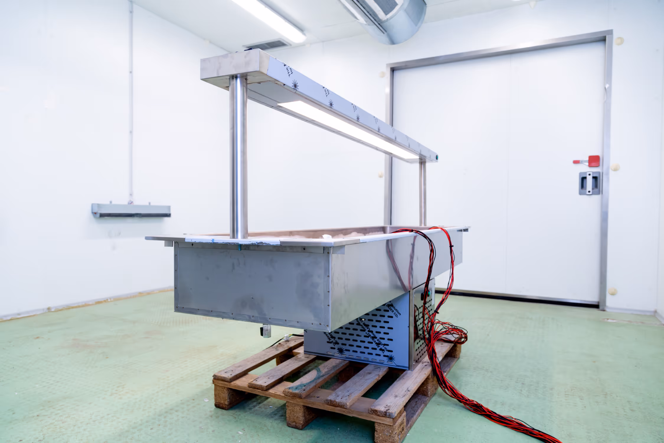 Large stainless steel industrial refrigeration unit with lighting, placed on a wooden pallet in a clean room with green floor and white walls.