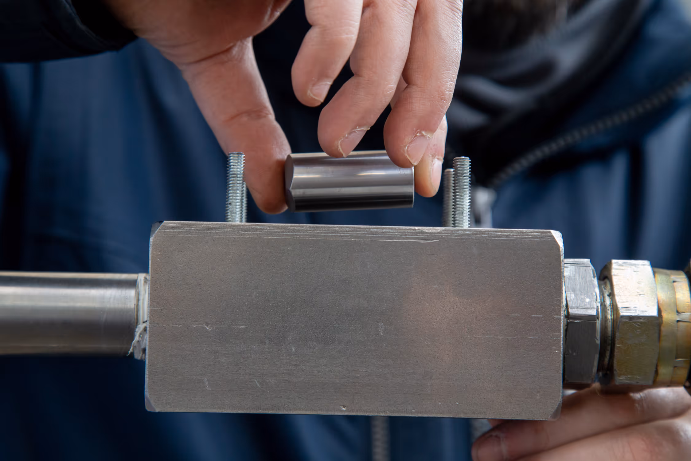 Close-up of a person’s hand holding a small cylindrical metal part above a rectangular metal block with two bolts.