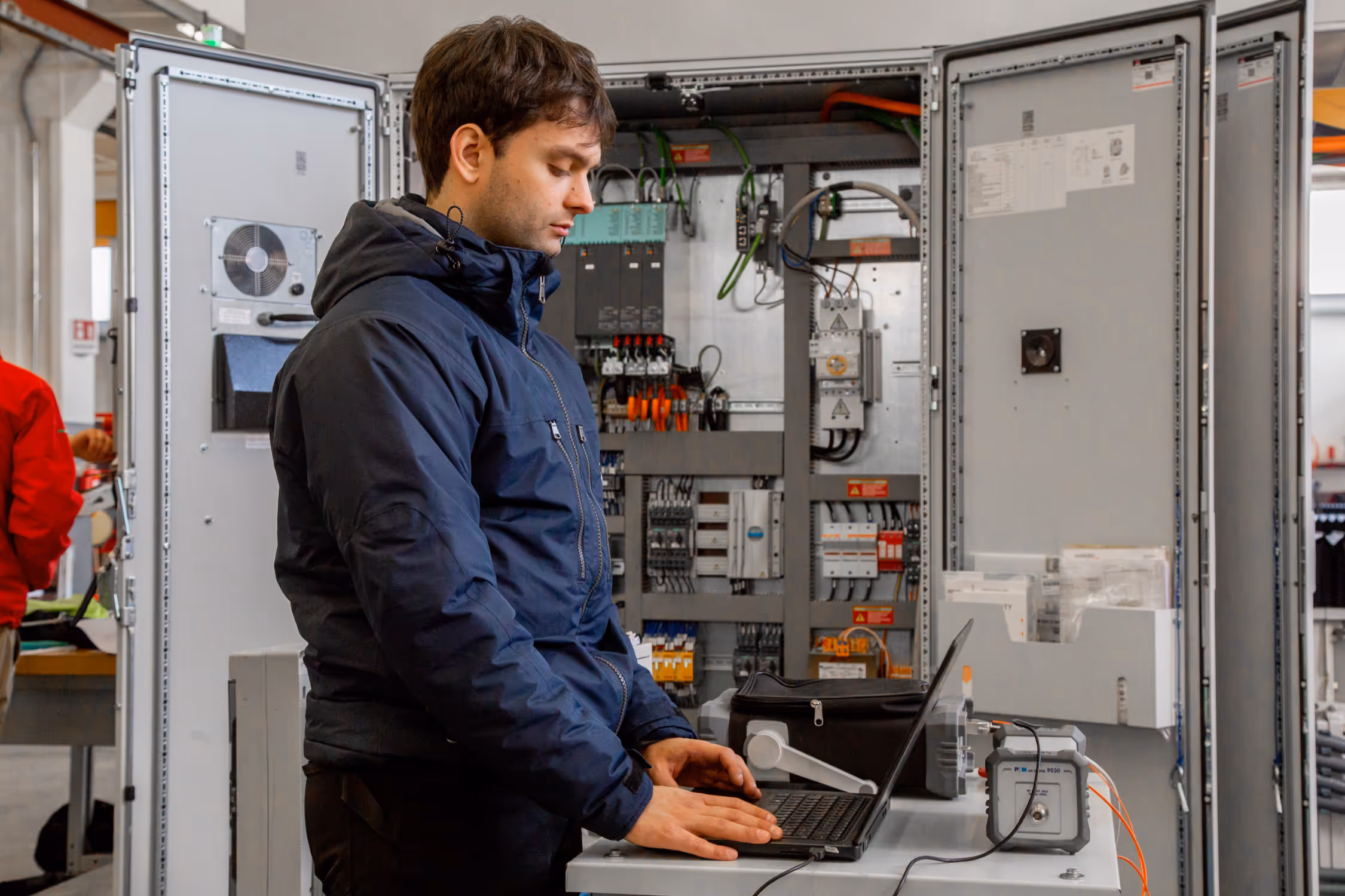 Technician working on a laptop in front of an open electrical control panel in an industrial setting.