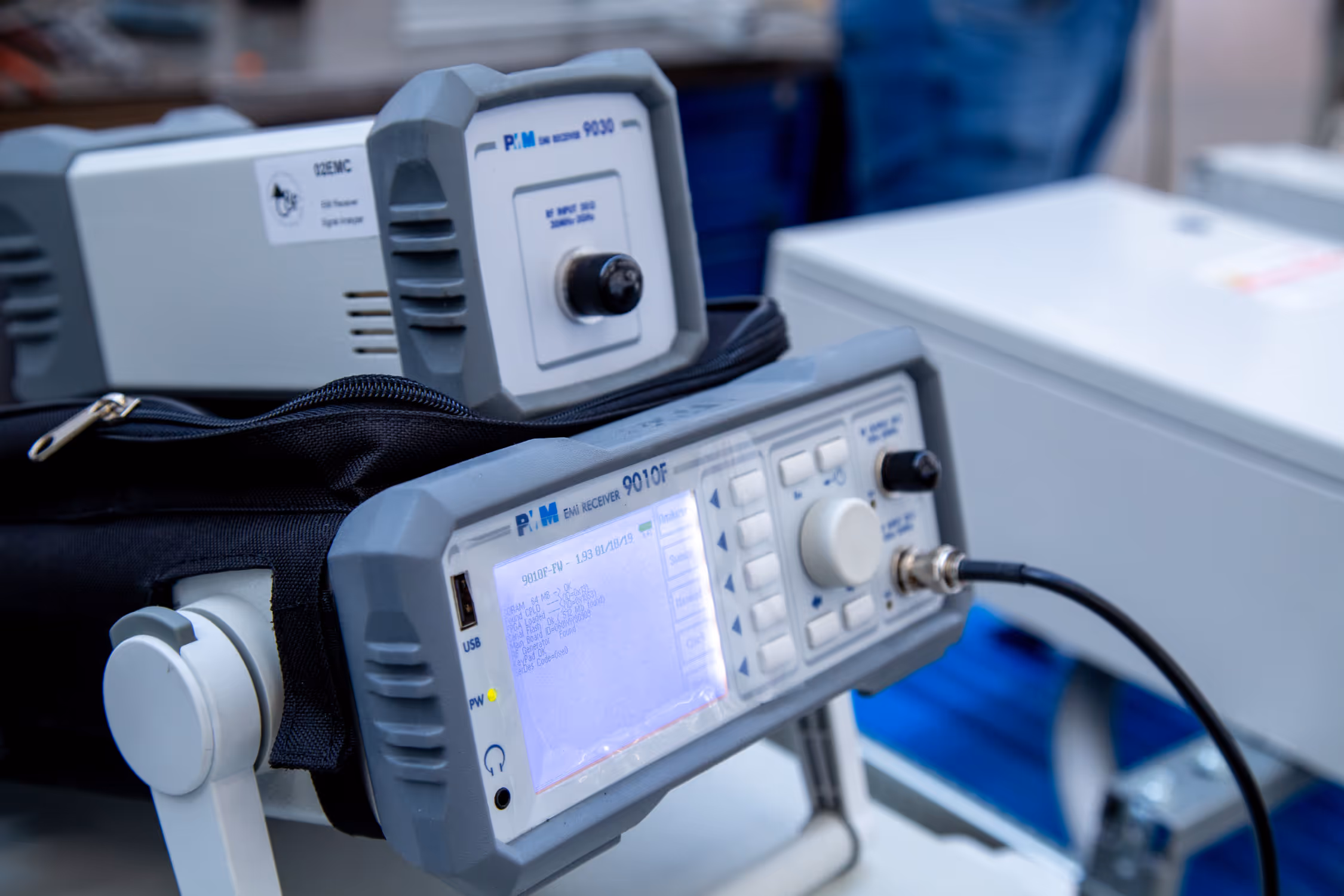 Close-up of a PM EMi receiver 9010F device with a glowing screen and control buttons, connected by a black cable.