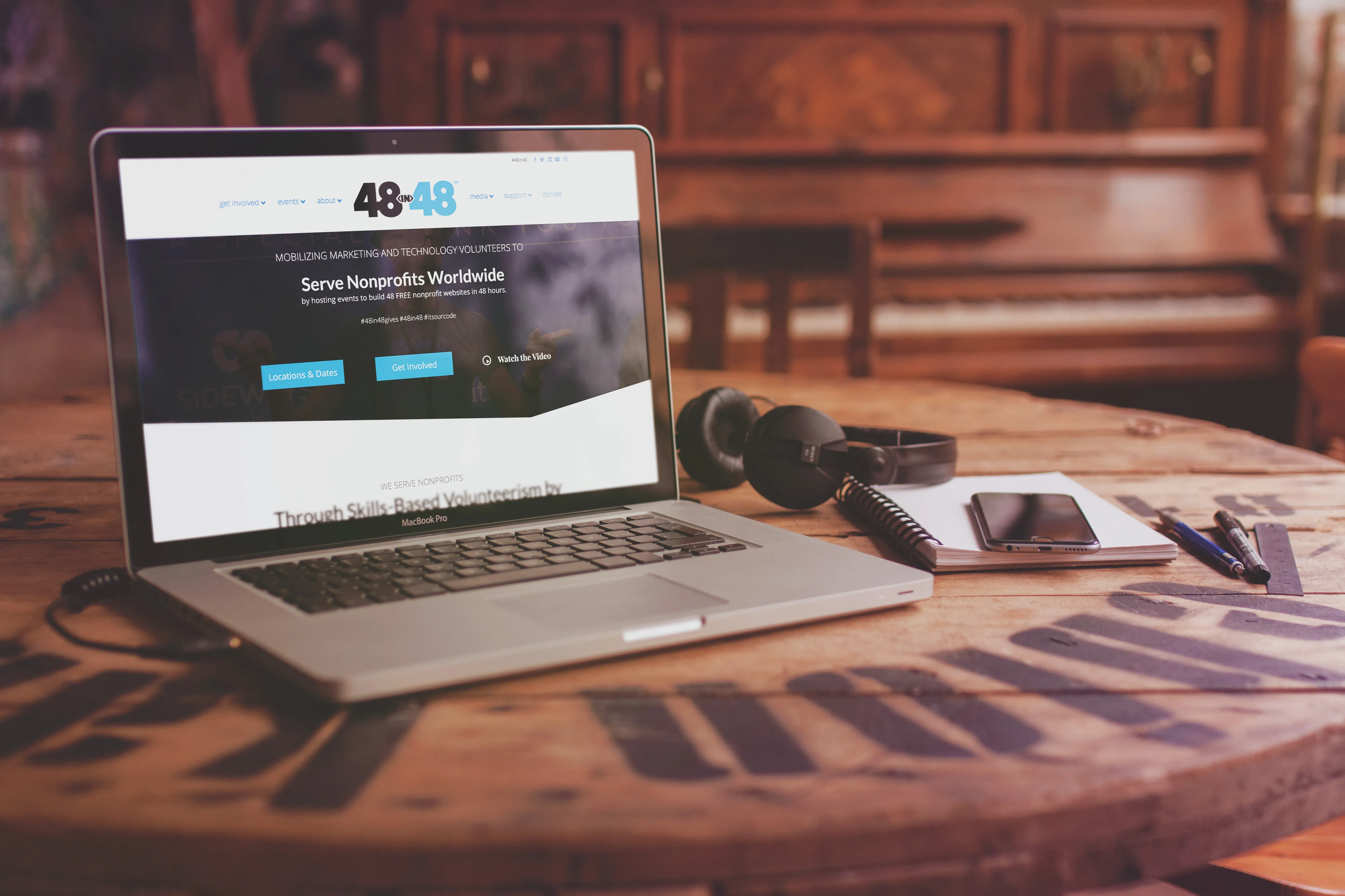 MacBook Pro laptop on a wooden table displaying a nonprofit website with headphones, notebook, smartphone, pens, and a ruler beside it.