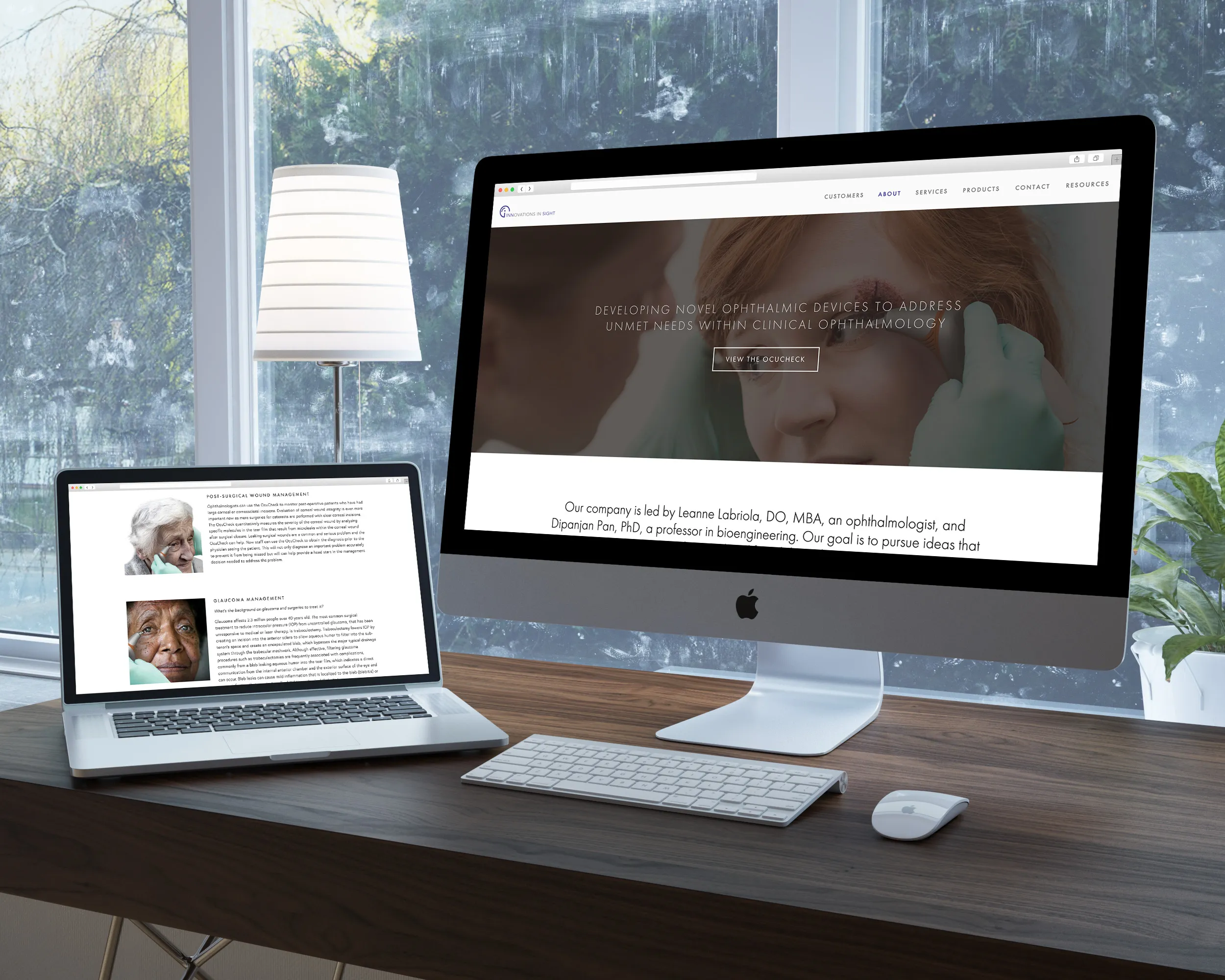 A modern workspace with a wooden desk featuring an iMac and a laptop displaying medical and ophthalmology-related content, next to a lamp and a plant by a window.
