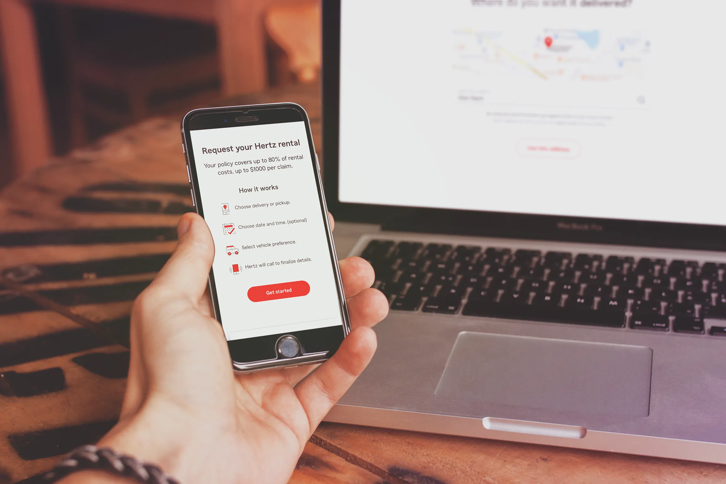 Hand holding a smartphone displaying a Hertz rental request screen with instructions and a red Get Started button, next to a laptop on a wooden table.