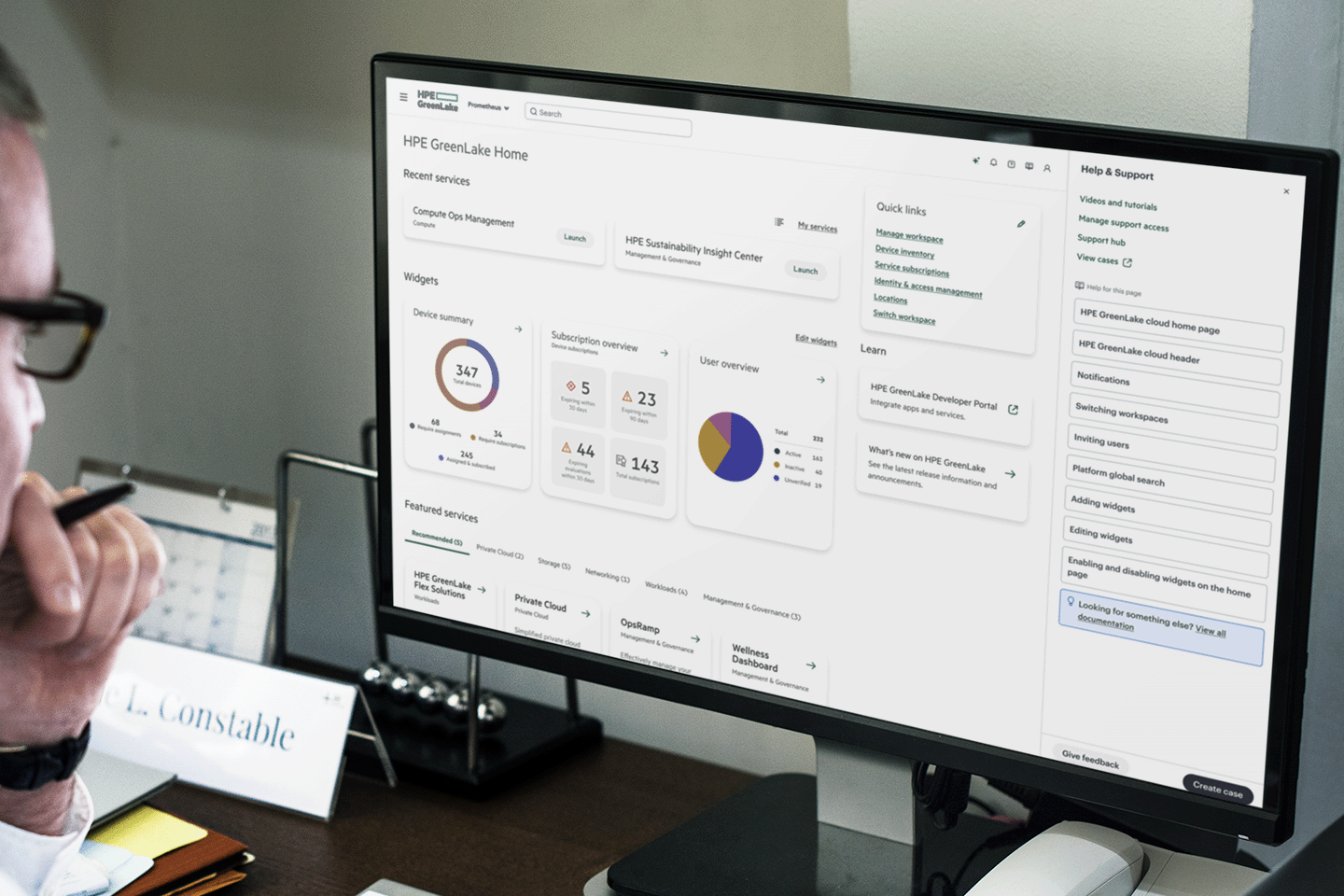 Computer monitor displaying the HPE GreenLake Home dashboard with charts, widgets, services, and help support options visible.