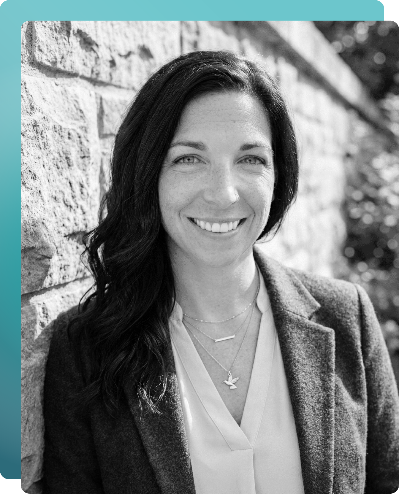 Smiling woman with long dark hair wearing layered necklaces and a blazer, standing against a stone wall.