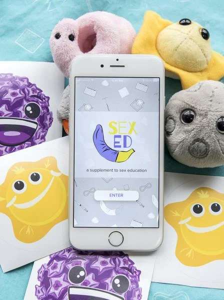 Smartphone displaying 'Sex Ed, a supplement to sex education' with an enter button, surrounded by colorful plush toys and cartoon character cards.