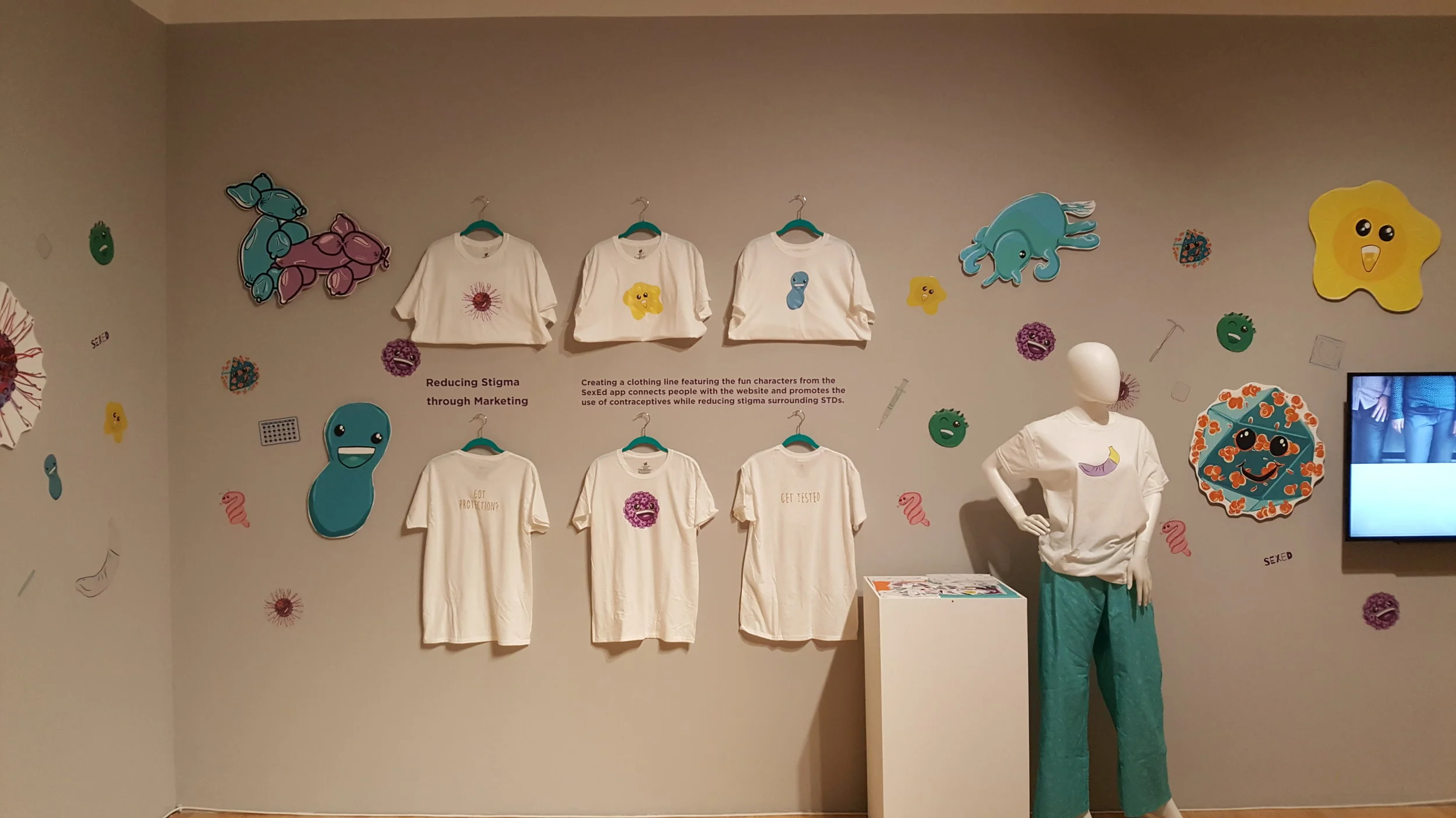 Exhibit wall displaying six white t-shirts with cartoon STD characters and messages like 'Get Tested' hung alongside colorful cartoon STD-themed artwork and a mannequin wearing a white t-shirt and teal pants.