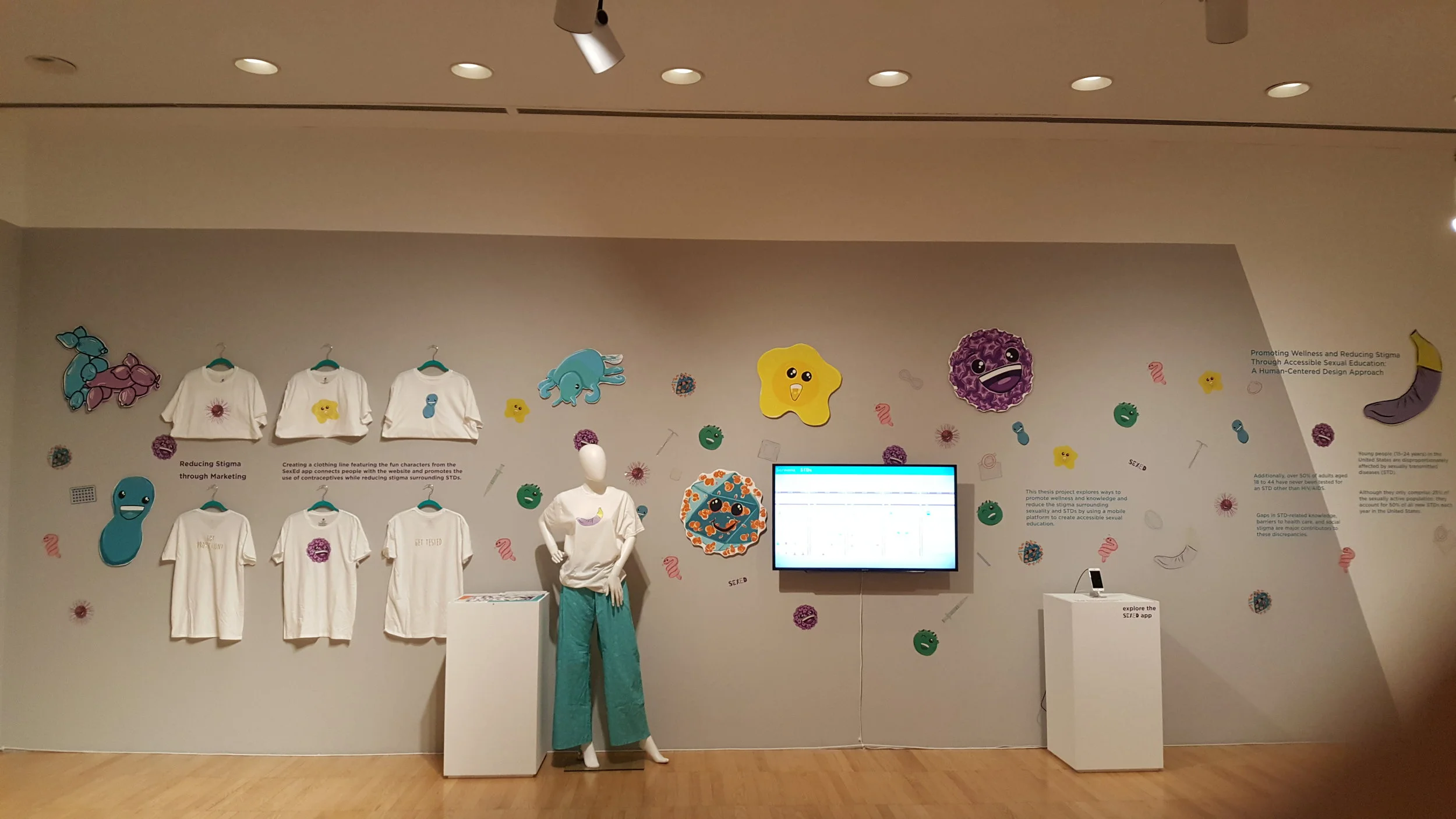 Exhibition wall with T-shirts featuring cartoon characters, a mannequin wearing a banana-themed shirt, colorful microorganism illustrations, a screen displaying data, and text about reducing stigma through sexual education.