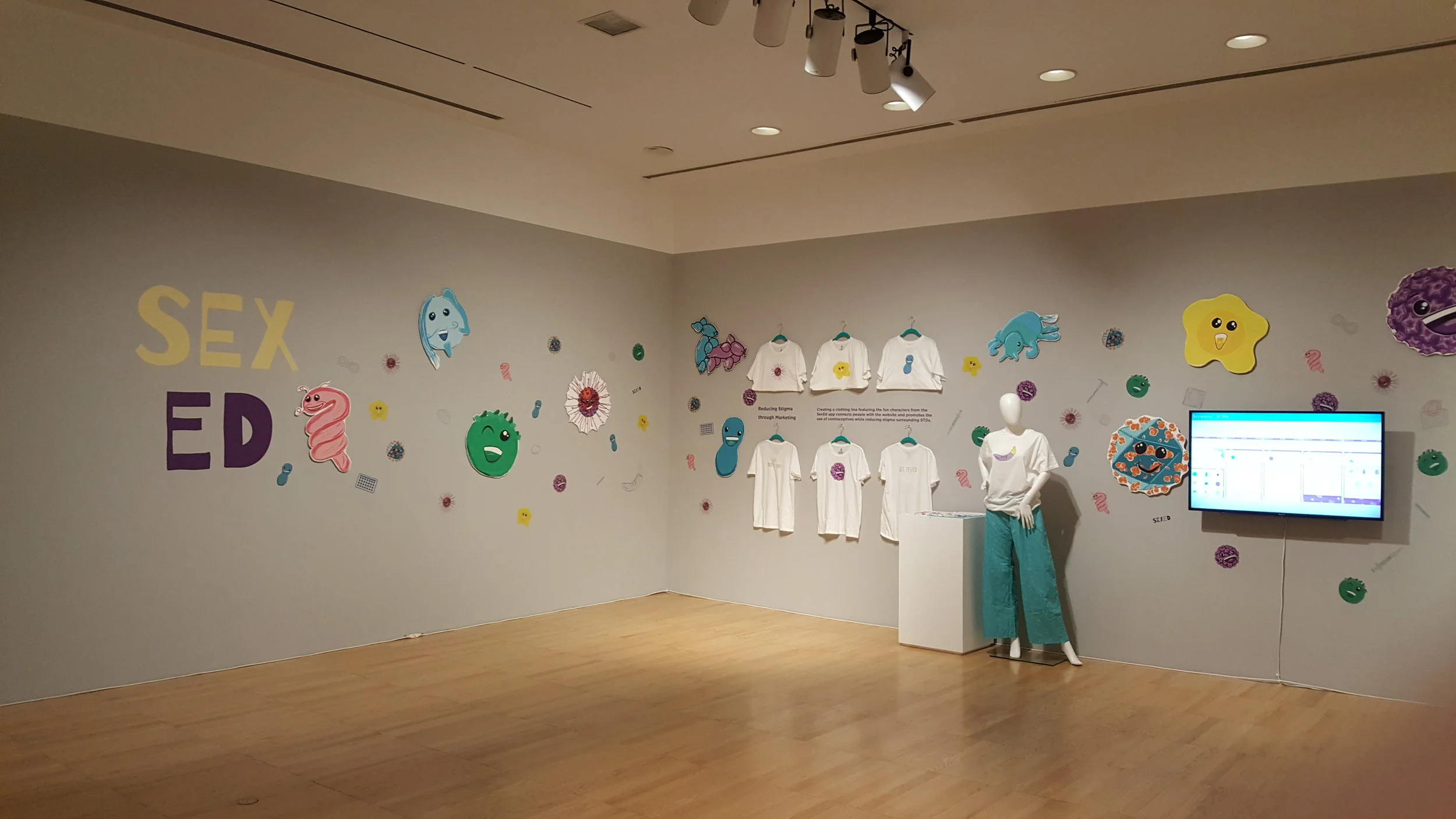 Exhibit room with wall text reading 'SEX ED' surrounded by colorful cartoon microbes, white t-shirts with microbe characters on hangers, a mannequin wearing a t-shirt and teal pants, and a screen displaying information.