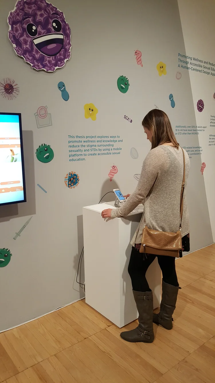 A woman interacts with a mobile device on a pedestal in a museum exhibit featuring colorful cartoon microbes and text about promoting wellness and accessible sexual education.