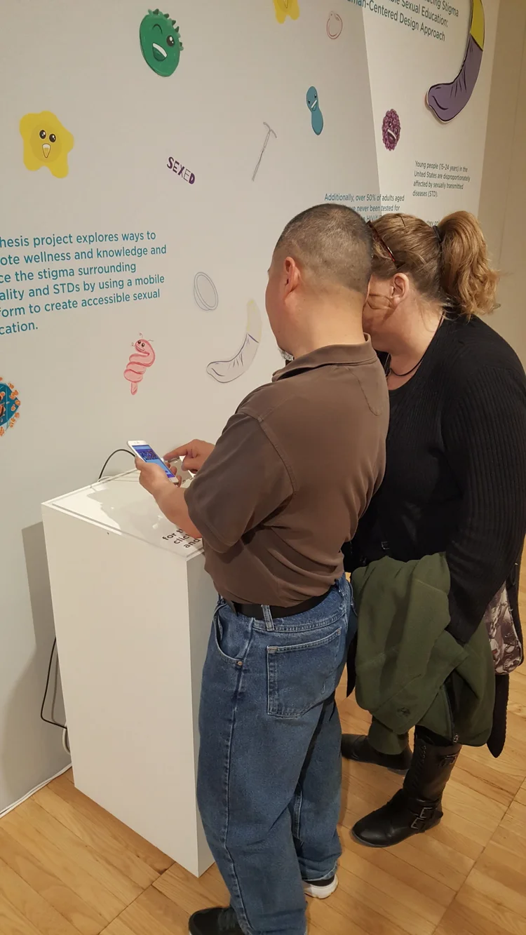 Two people interacting with a touchscreen device on a white pedestal, in front of a wall with colorful sexual health education illustrations and text.
