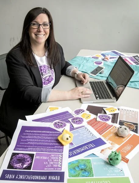 Smiling woman with glasses seated at a table with a laptop, surrounded by colorful educational posters and small plush toys representing viruses.