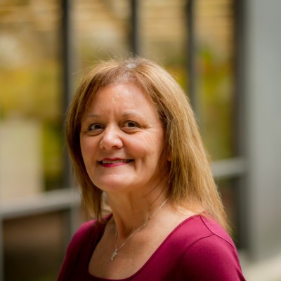 Smiling middle-aged woman with shoulder-length light brown hair wearing a maroon top and a necklace in an indoor setting with blurred background.