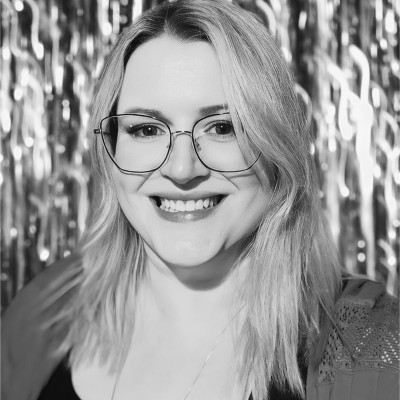 Smiling woman with shoulder-length hair and glasses, standing against a shiny, ridged backdrop.