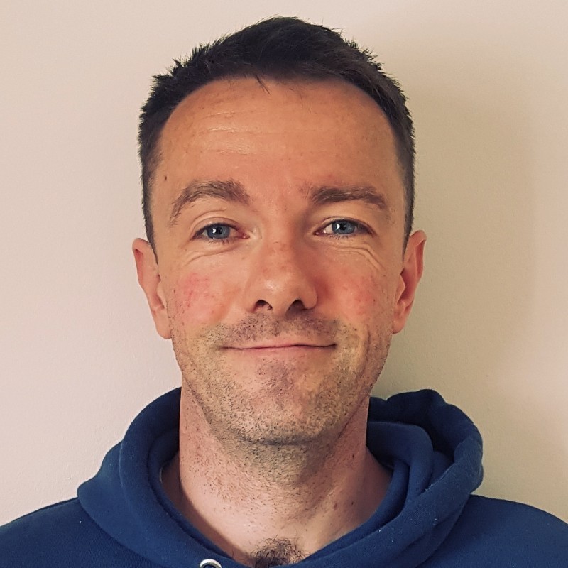 Smiling man with short dark hair and blue eyes wearing a dark blue hoodie against a light wall.