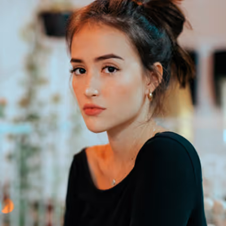 Young woman with dark hair tied back wearing a black top, looking thoughtfully at the camera.