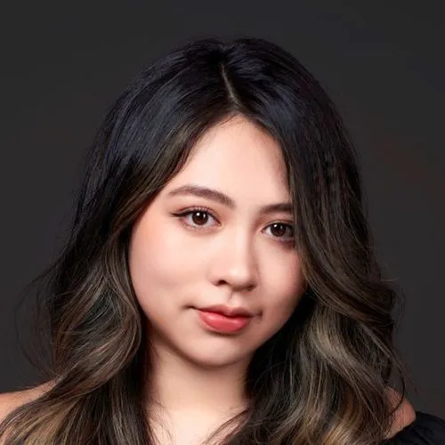 Close-up portrait of a young woman with long dark wavy hair and natural makeup looking at the camera against a dark background.