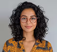 Smiling woman with curly hair and round glasses wearing a yellow patterned top against a gray background.