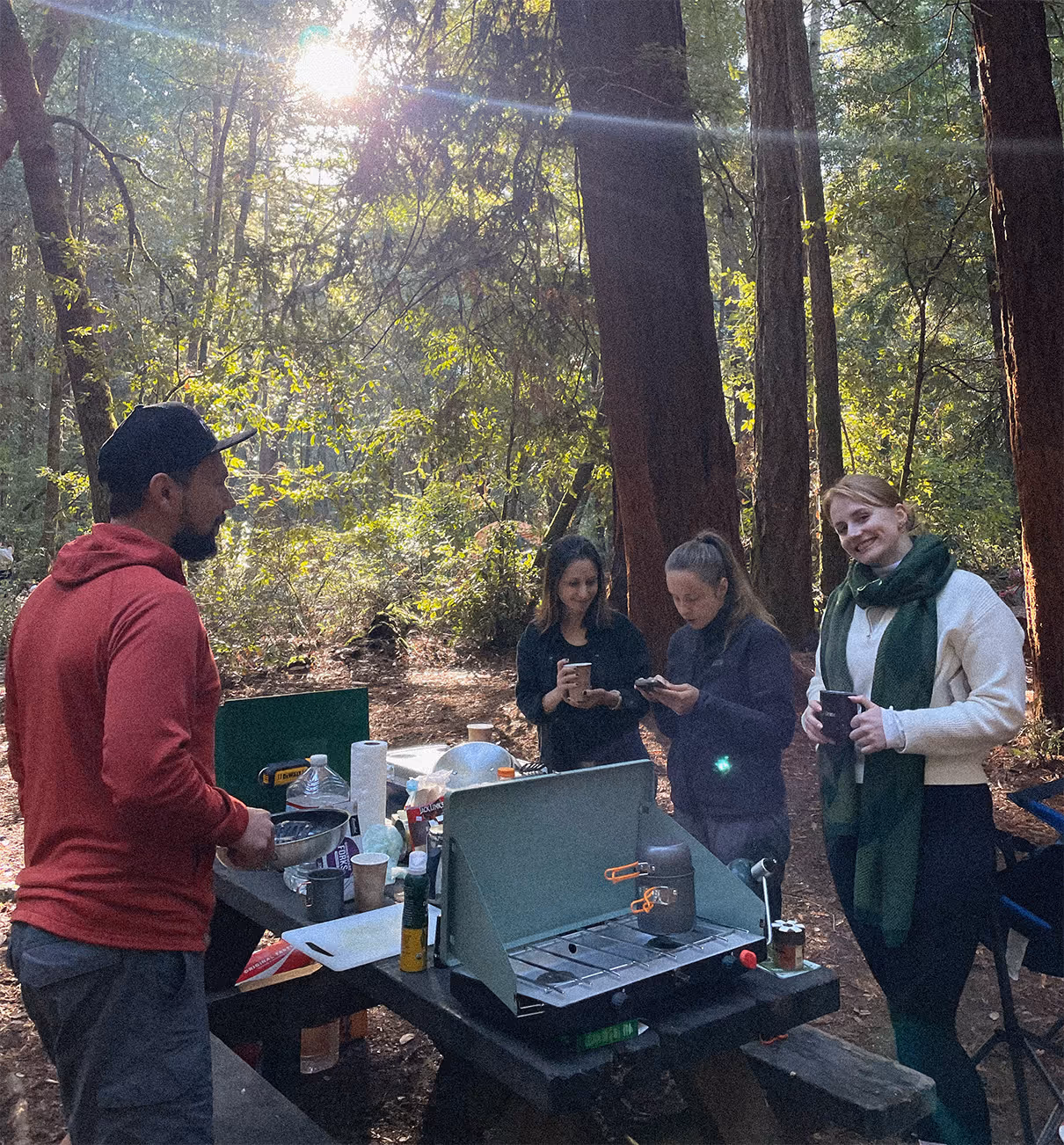 Four people gathered around a picnic table with camping gear and a stove in a sunlit forest.