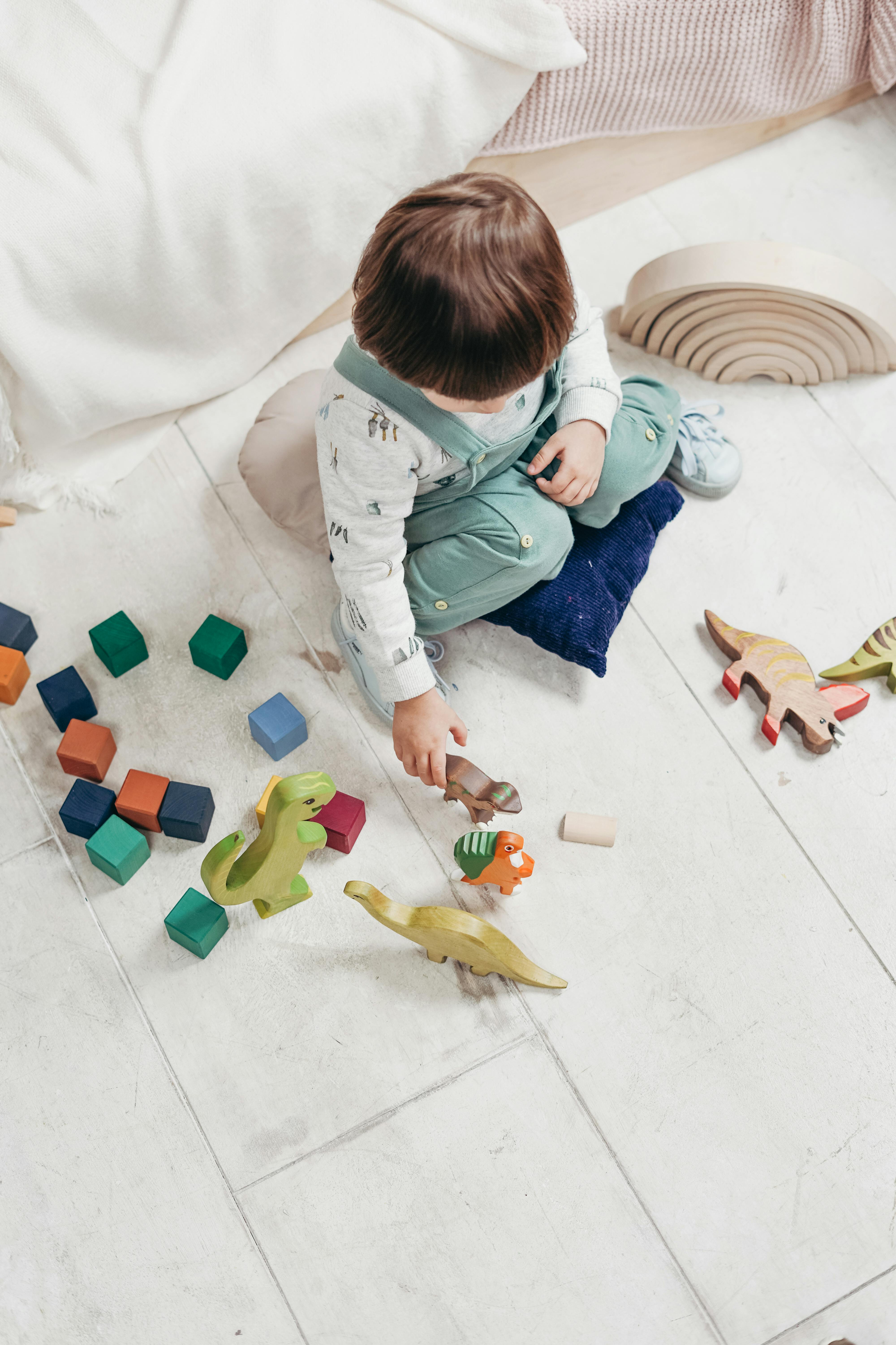 Top view of toddler sitting on cushions playing with wooden dinosaur toys and colorful blocks on a light tile floor.