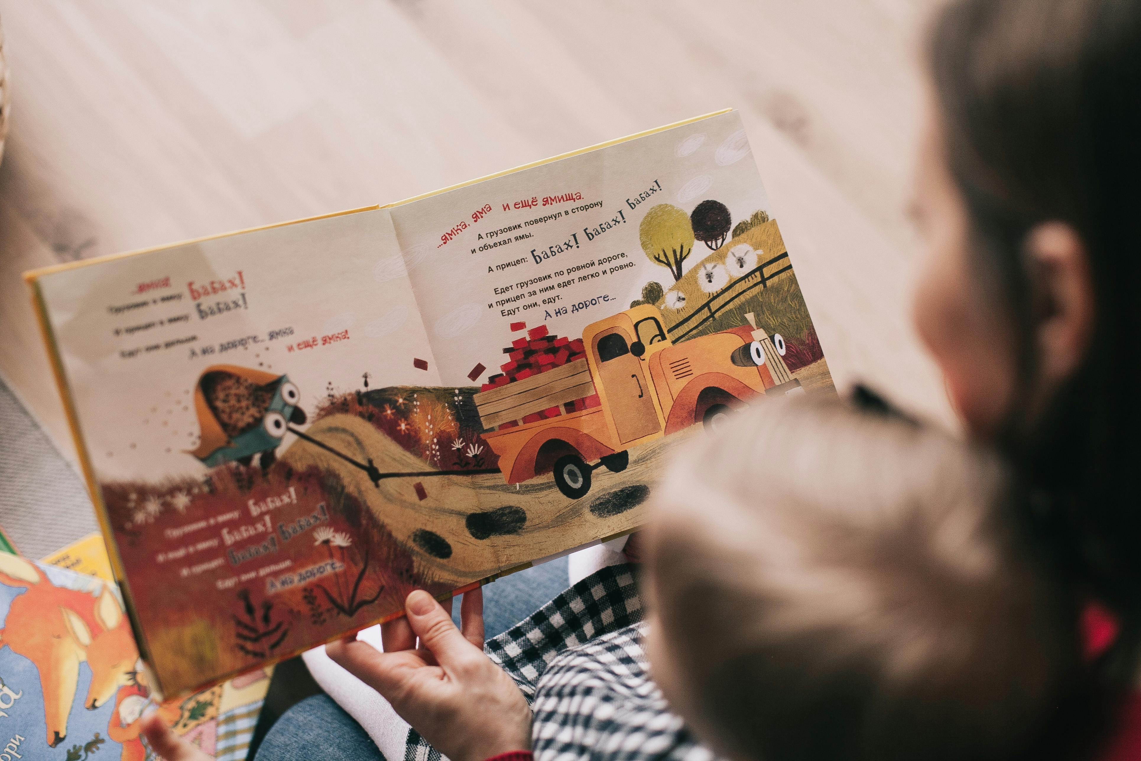 Adult reading a colorful children’s book with a cartoon truck and owl illustrations to a child.