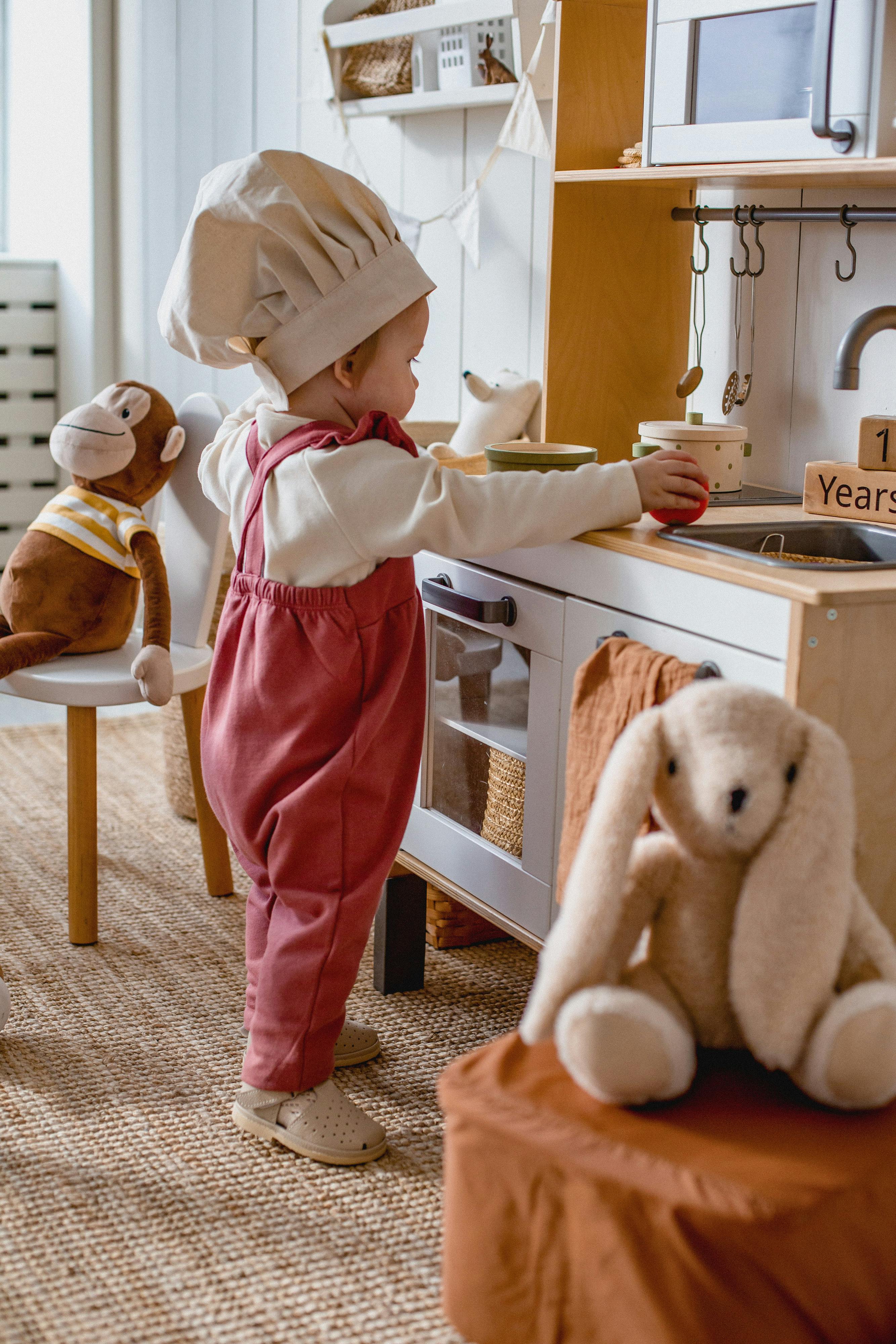Toddler wearing a chef hat and red overalls playing with a toy kitchen set, surrounded by stuffed animals.