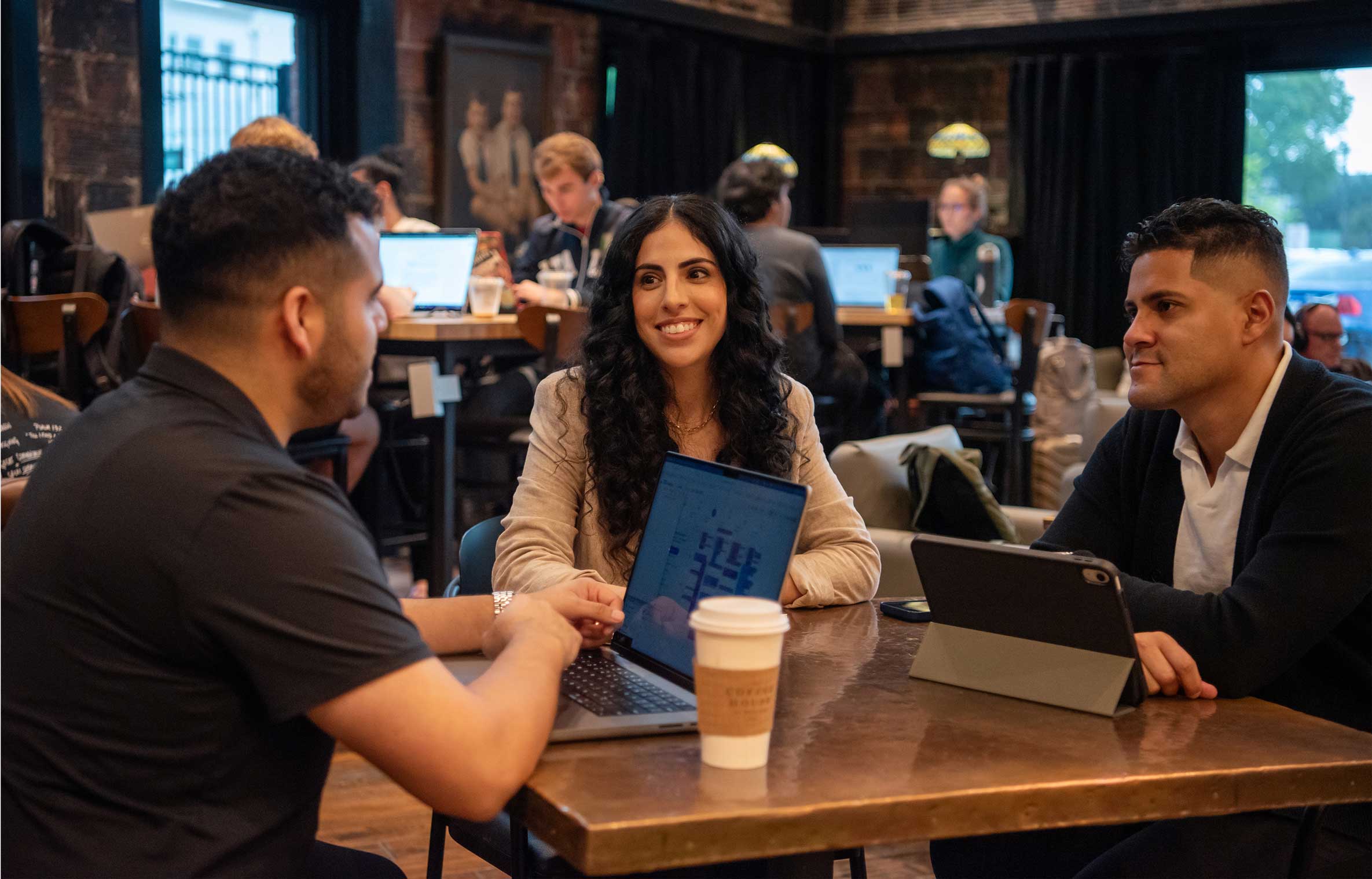 Three people sitting at a table with a laptop, tablet, and coffee cup, engaged in conversation in a cozy cafe.