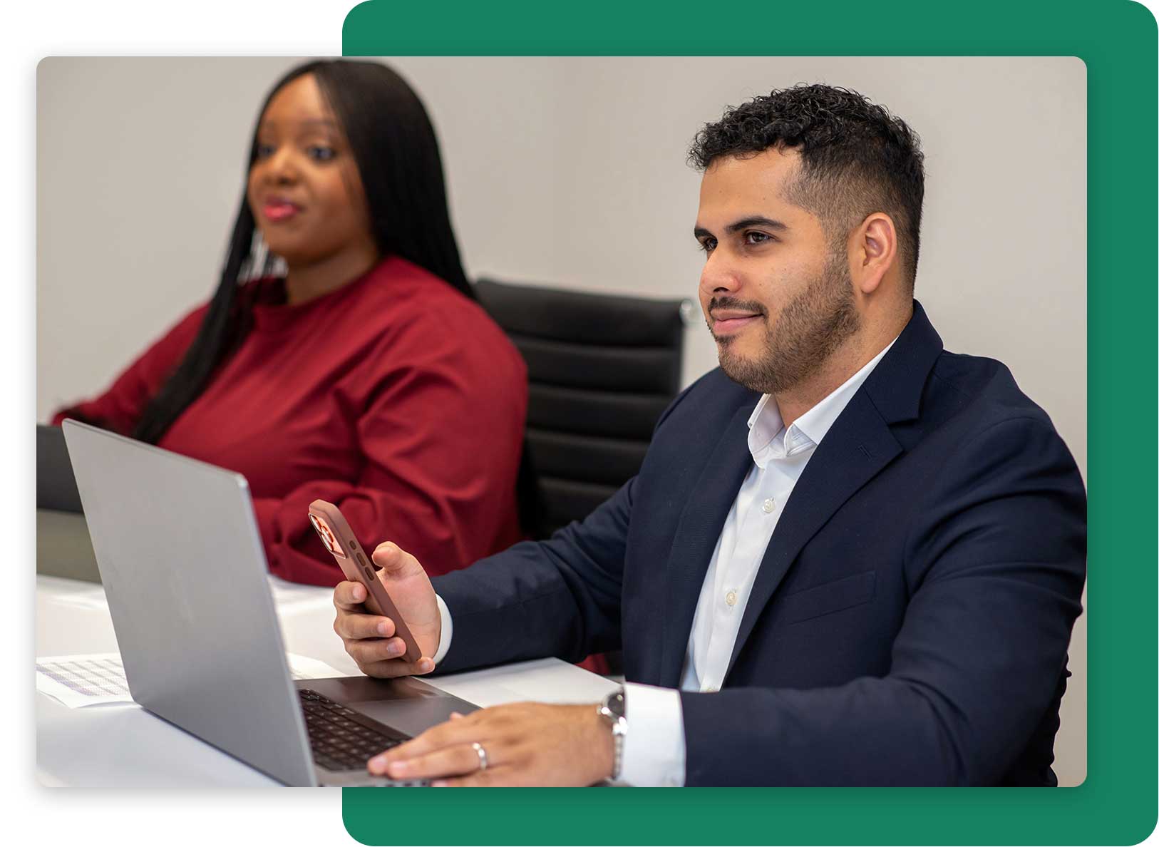 Two business professionals, a man in a suit using a smartphone and laptop, and a woman in a red blouse sitting beside him.