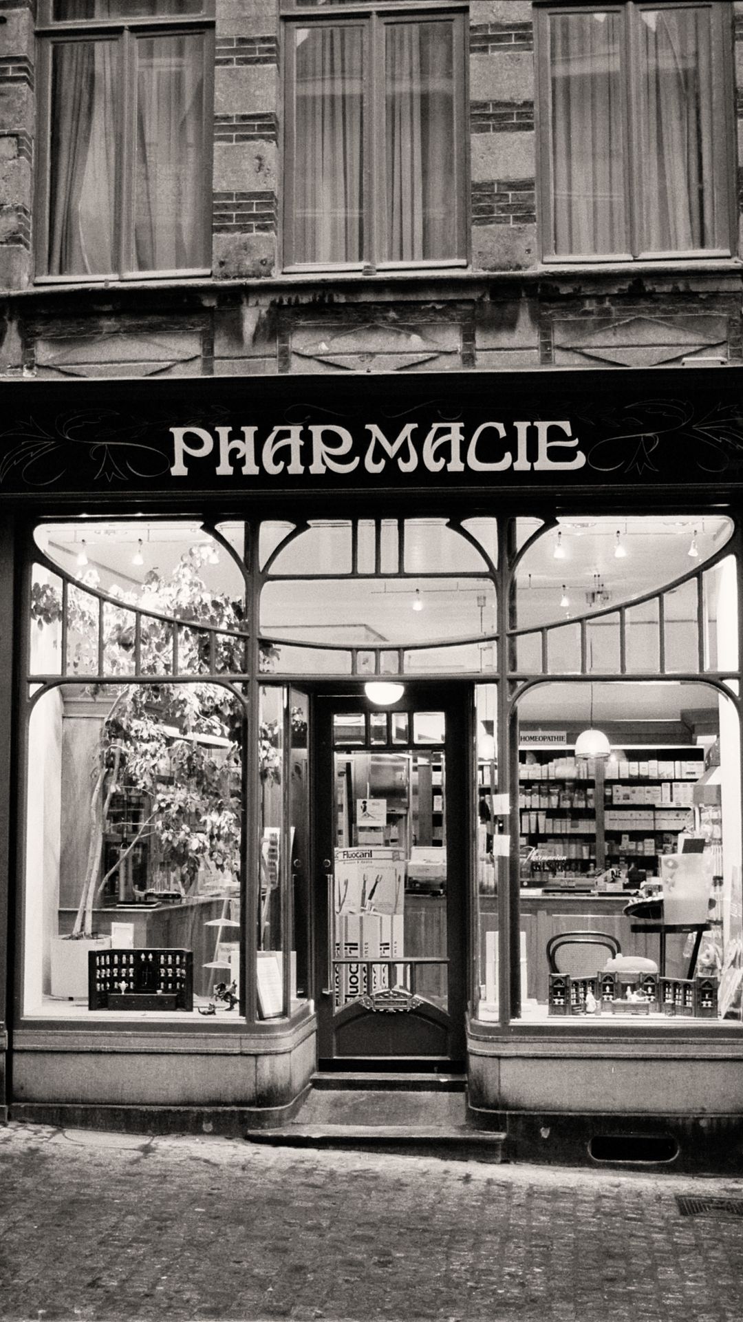 Façade ancienne d'une officine en noir et blanc, enseigne Pharmacie au-dessus d'une vitrine en bois et d'une rue pavée