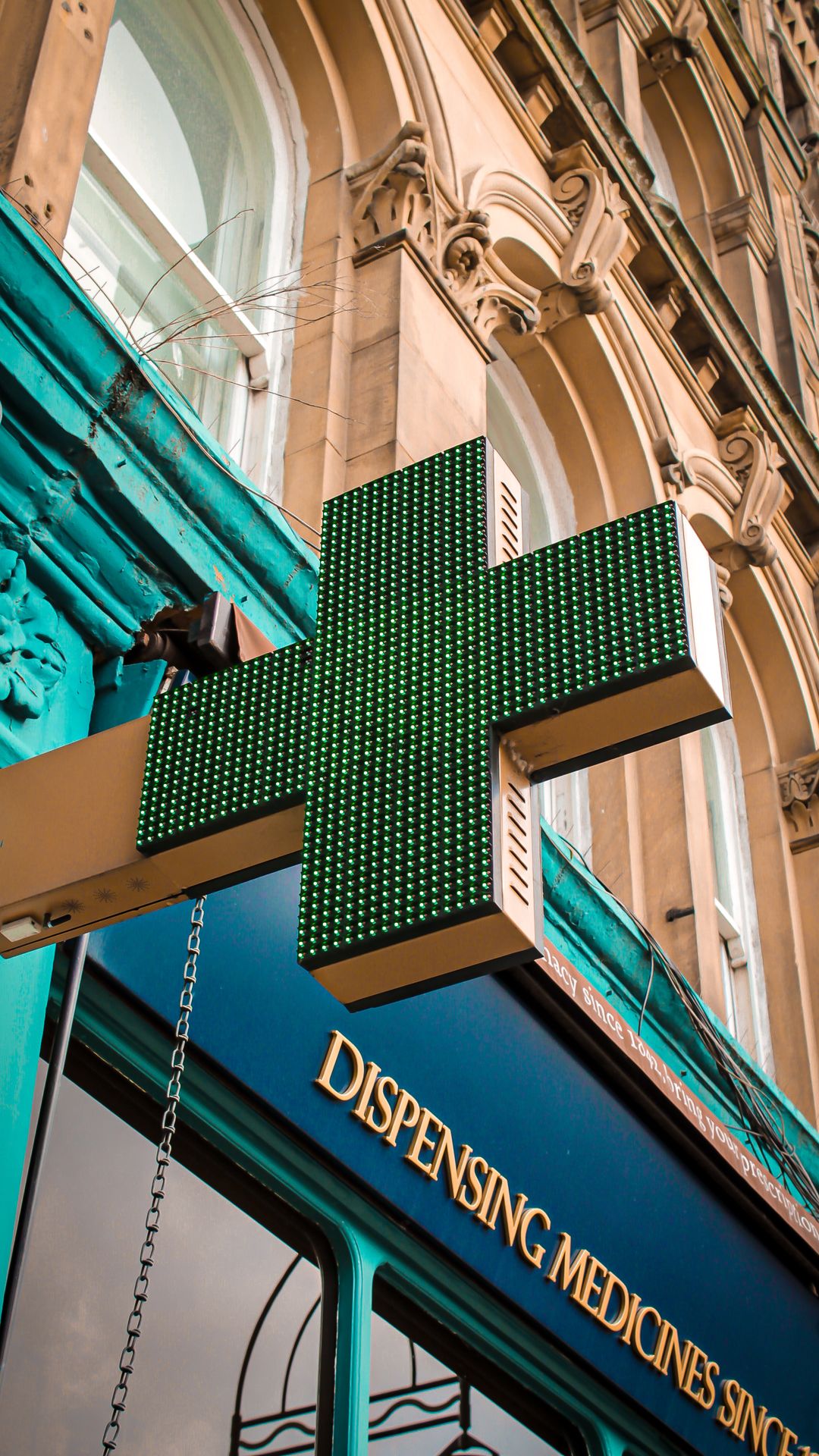 Croix verte lumineuse de pharmacie fixée à une façade ancienne, au-dessus d'une devanture bleue à lettres dorées