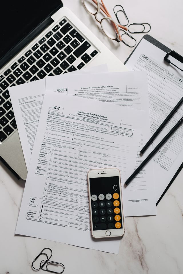 Formulaires fiscaux posés sur un bureau en marbre avec ordinateur portable, calculatrice de téléphone et trombones