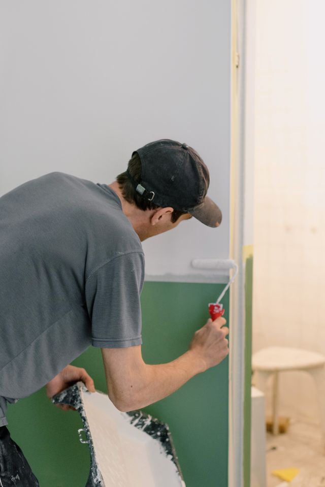 Ouvrier en casquette peignant au rouleau l'encadrement d'une porte dans un appartement en travaux